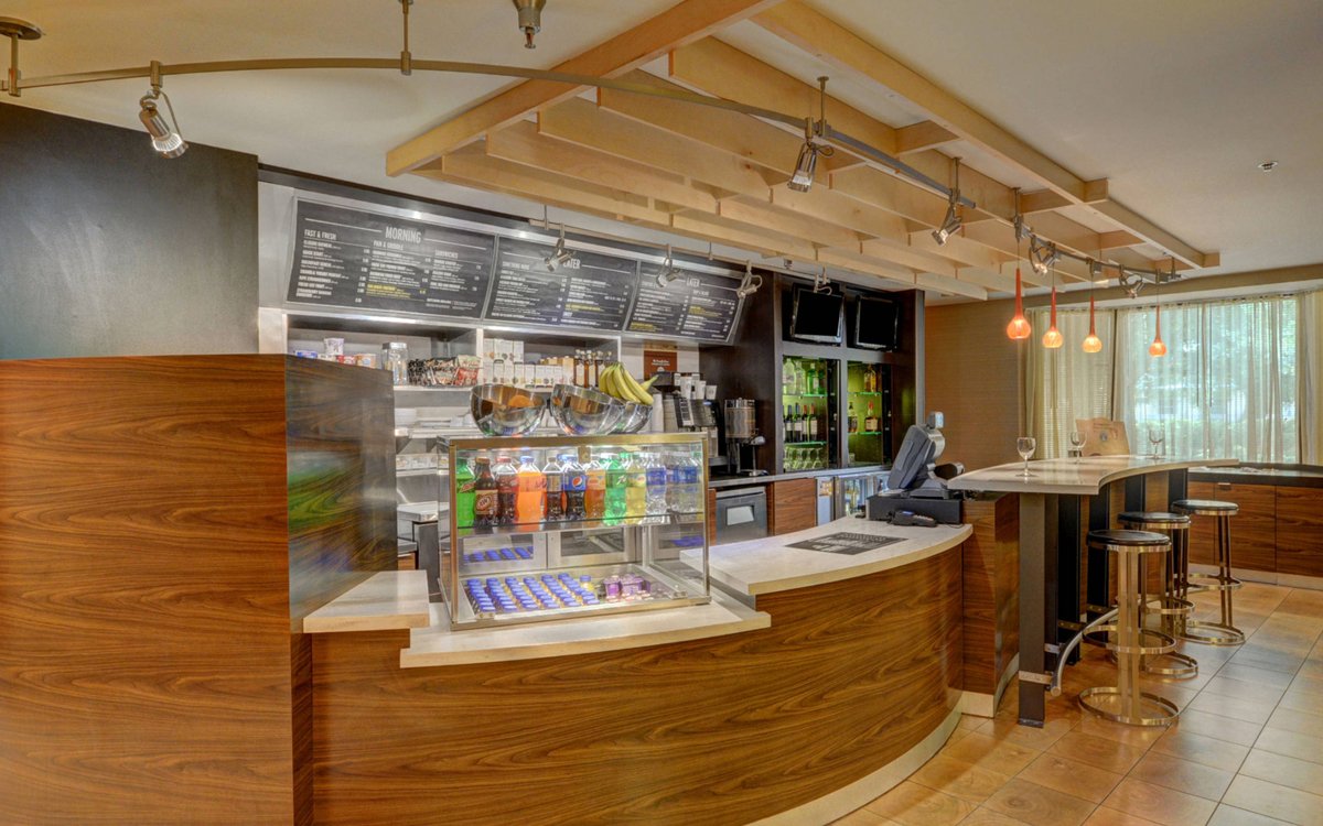 Interior of a modern coffee shop or cafe with a curved wooden counter, a glass display with beverages, a cash register, and a menu board overhead. There are bar stools and large windows with curtains allowing natural light.
