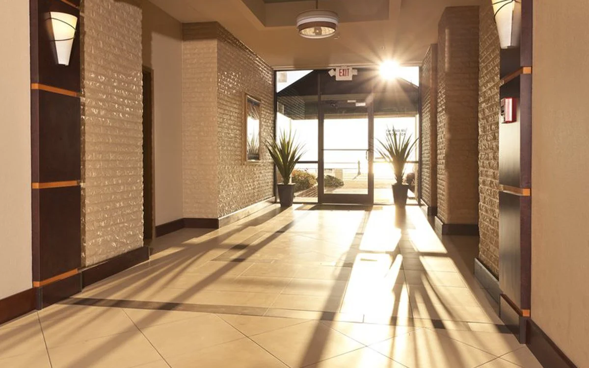 Inside of a well-lit building lobby with tile flooring, brick walls, potted plants near the glass door, sunlight streaming through, and an exit sign above the door.