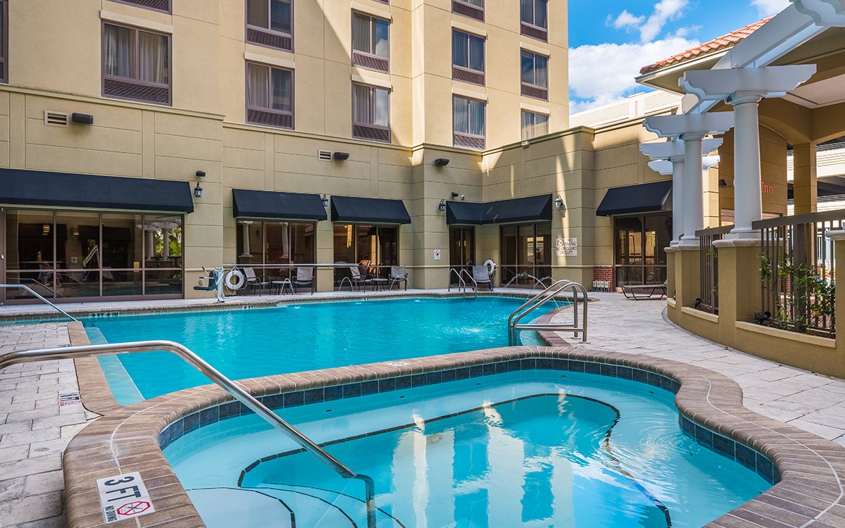 Outdoor hotel swimming pool and hot tub area with buildings and blue sky in the background.