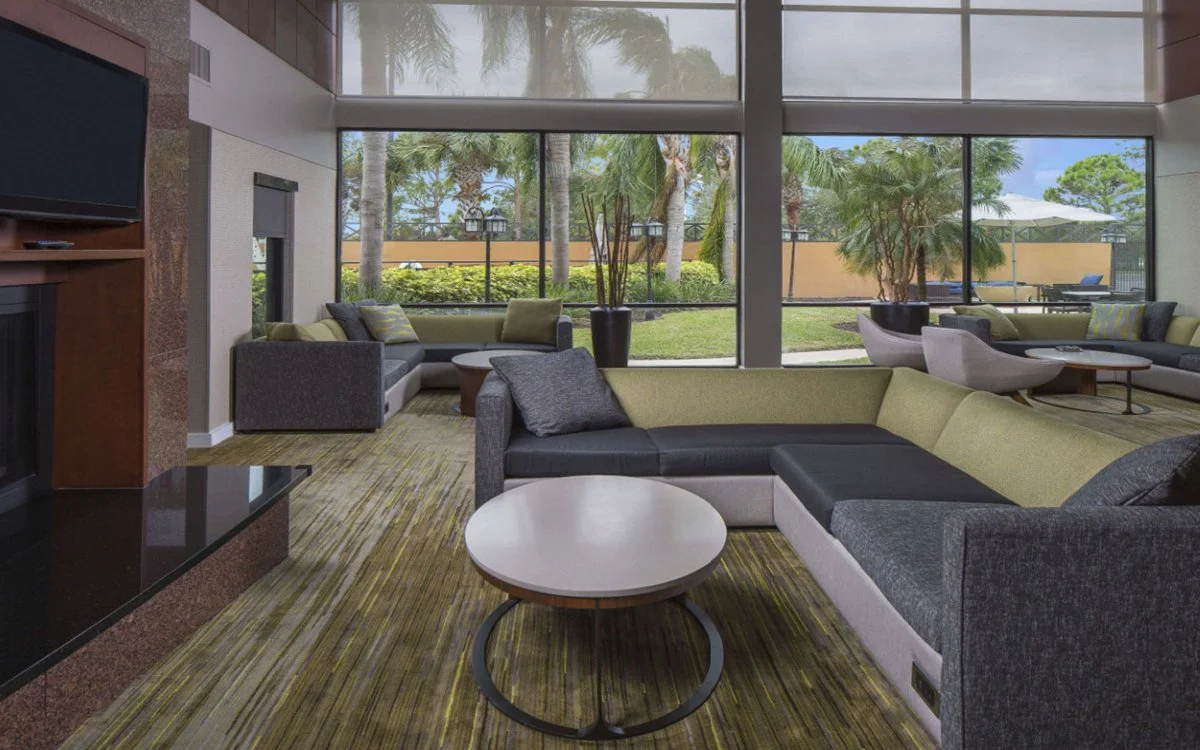 Modern living room with large windows, tropical plants outside, and contemporary furniture including L-shaped sofa, armchairs, and coffee tables.