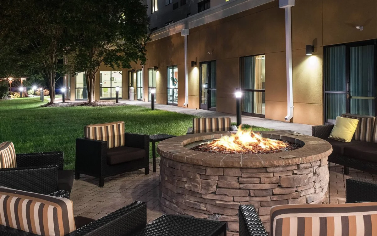 Outdoor patio area with a lit fire pit, surrounded by chairs with striped cushions, in front of a building with large glass windows at night.