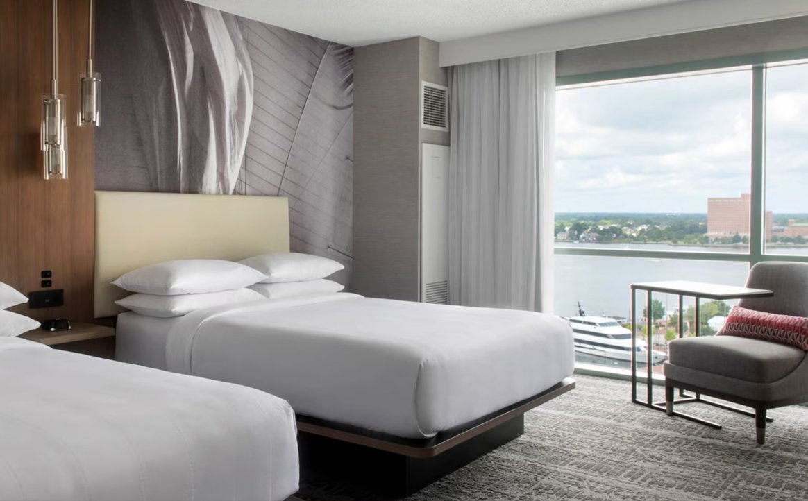 A hotel room with two beds, a large window overlooking a river and cityscape, a chair with a side table, and wall art behind one bed.