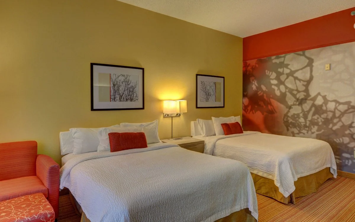 Hotel room with two beds, a beige wall, framed artwork, a lamp, and a patterned wall with shadows.