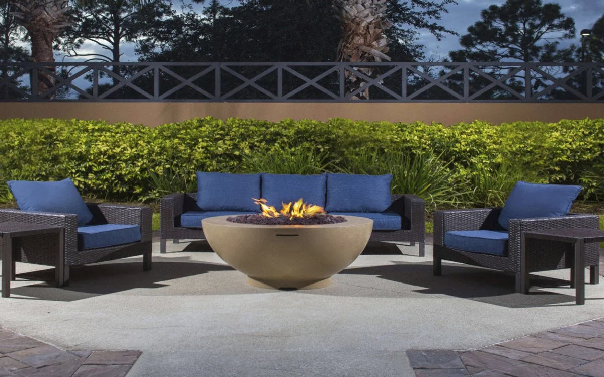 Outdoor patio with a fire pit table, surrounded by three blue-cushioned wicker chairs, with yellow-green bushes and trees in the background.