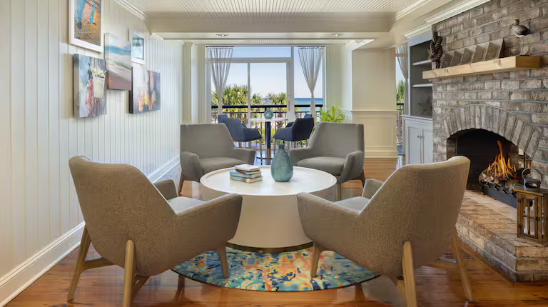 Living room with four beige armchairs around a white round coffee table on a colorful rug, fireplace on the right, artwork on the left wall, large windows with curtains, overlooking a balcony with ocean view.