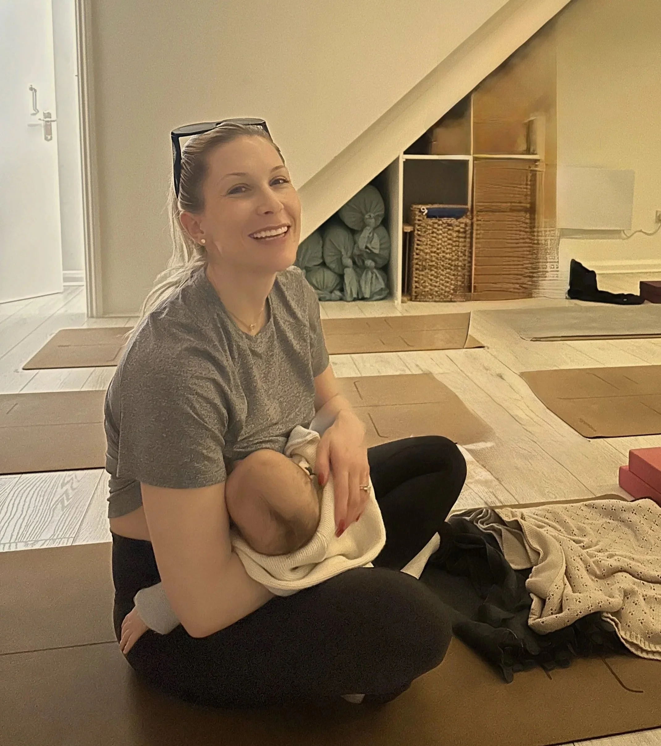 A smiling woman sitting on a yoga mat while breastfeeding a baby, with a cozy, organized room in the background.