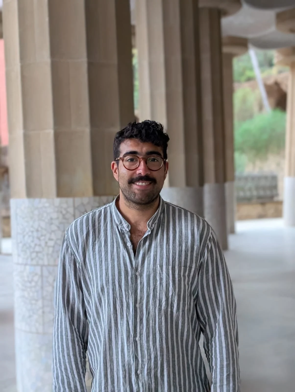 A man with dark hair, mustache, and glasses wearing a striped button-up shirt, standing outdoors near beige stone columns and a building with a modern architectural design.