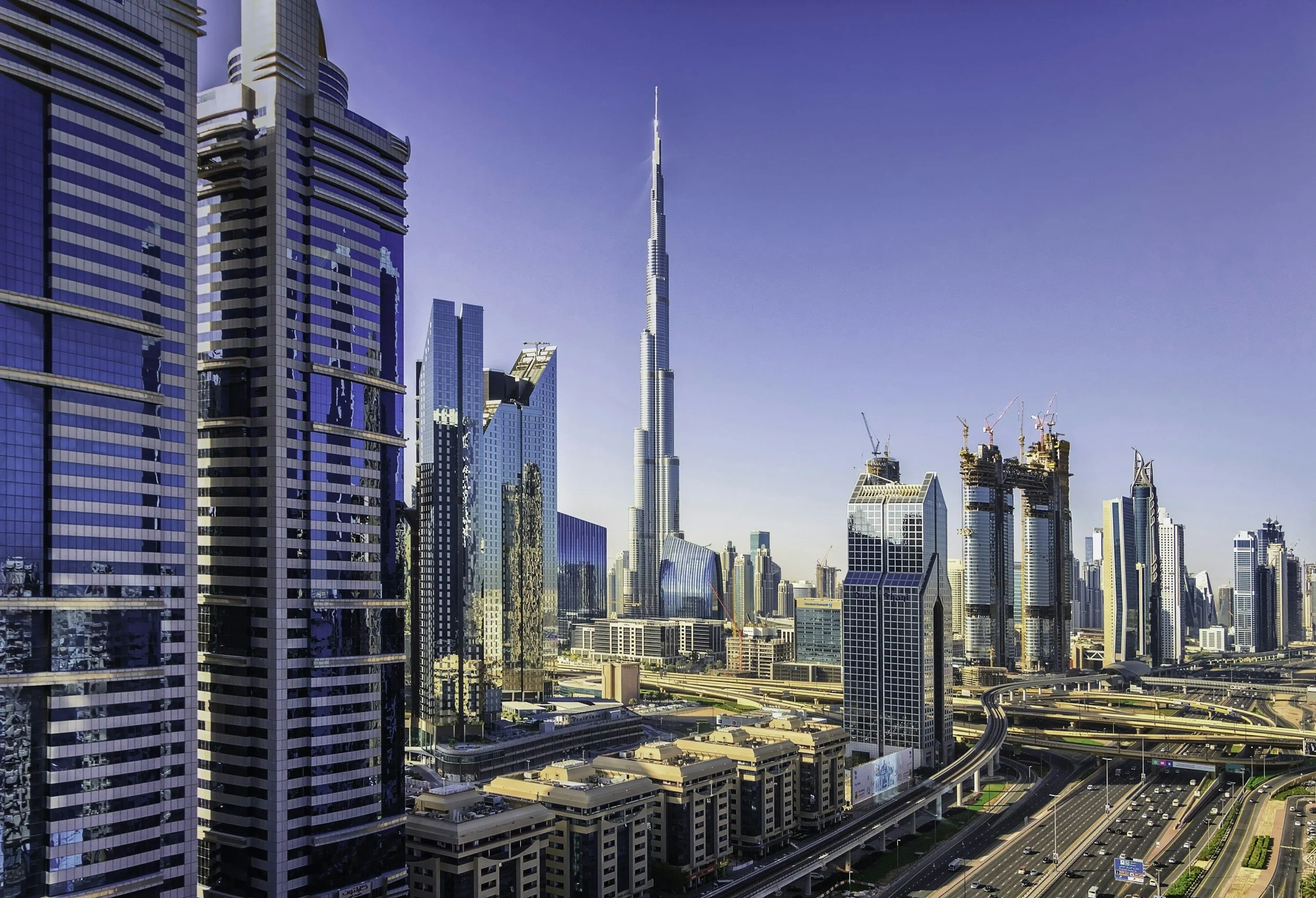 Vista aérea del centro de Dubái con rascacielos modernos, incluyendo la Torre Burj Khalifa en el centro y edificios en construcción bajo un cielo despejado.