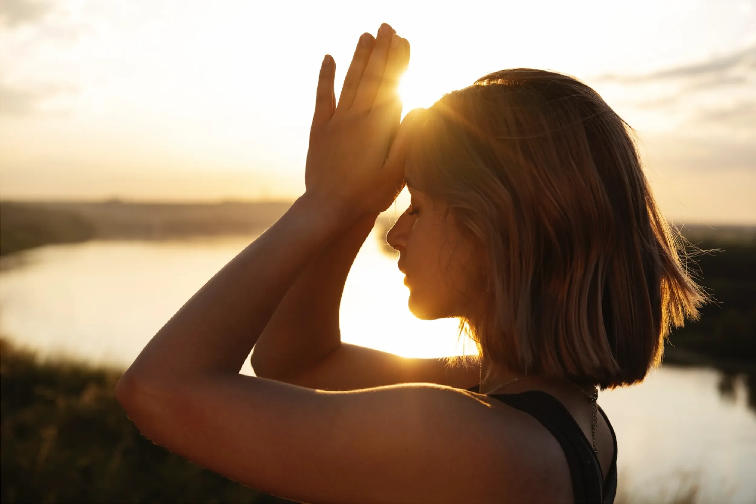 Young woman in profile with hands pressed together in prayer against her forehead at golden sunset by a river, representing Universal Human Foundation's support for mindfulness, gratitude, and inner reflection.