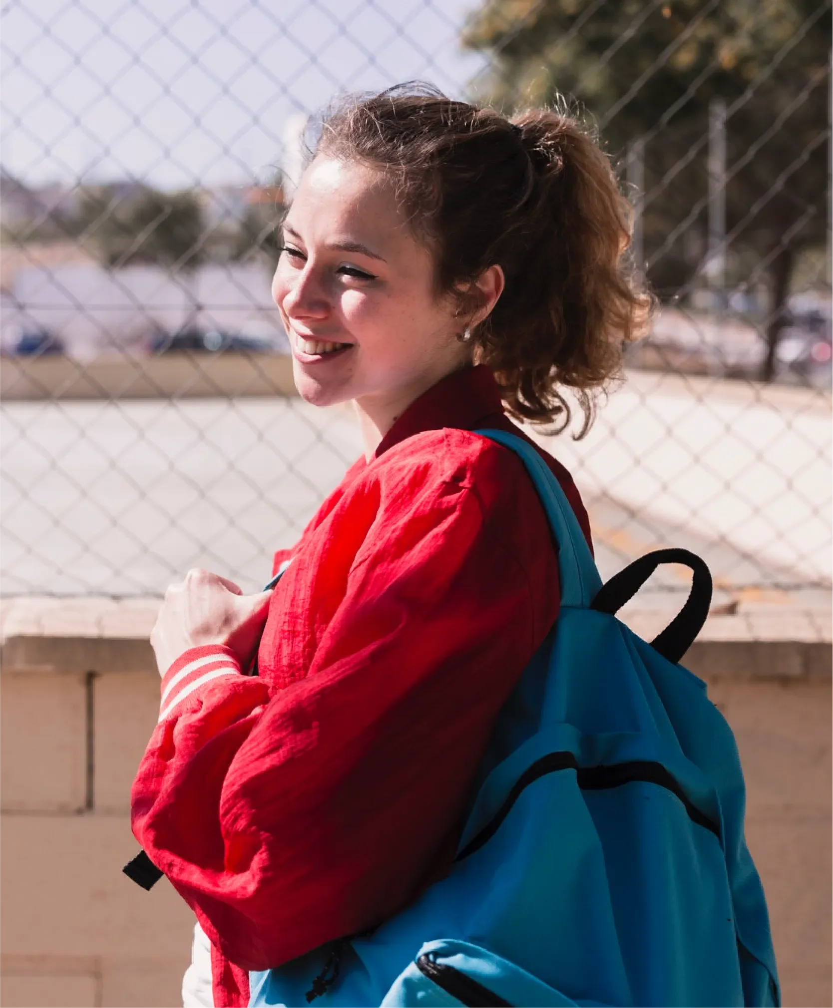 Young student with a red jacket and blue backpack smiling over her shoulder outdoors, representing Universal Human Foundation's youth programs supporting personal growth and wellbeing.