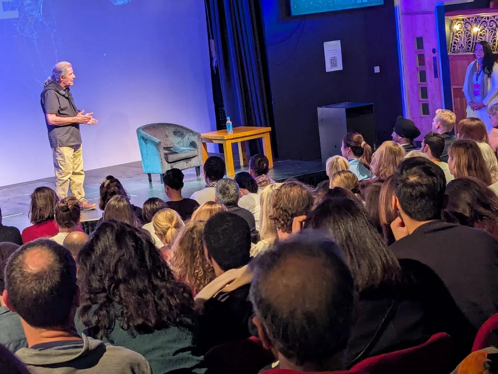 Gary Zukav speaking on stage to a seated audience in an intimate event setting, with a chair and table beside him, creating a space for reflection, learning, and shared spiritual exploration