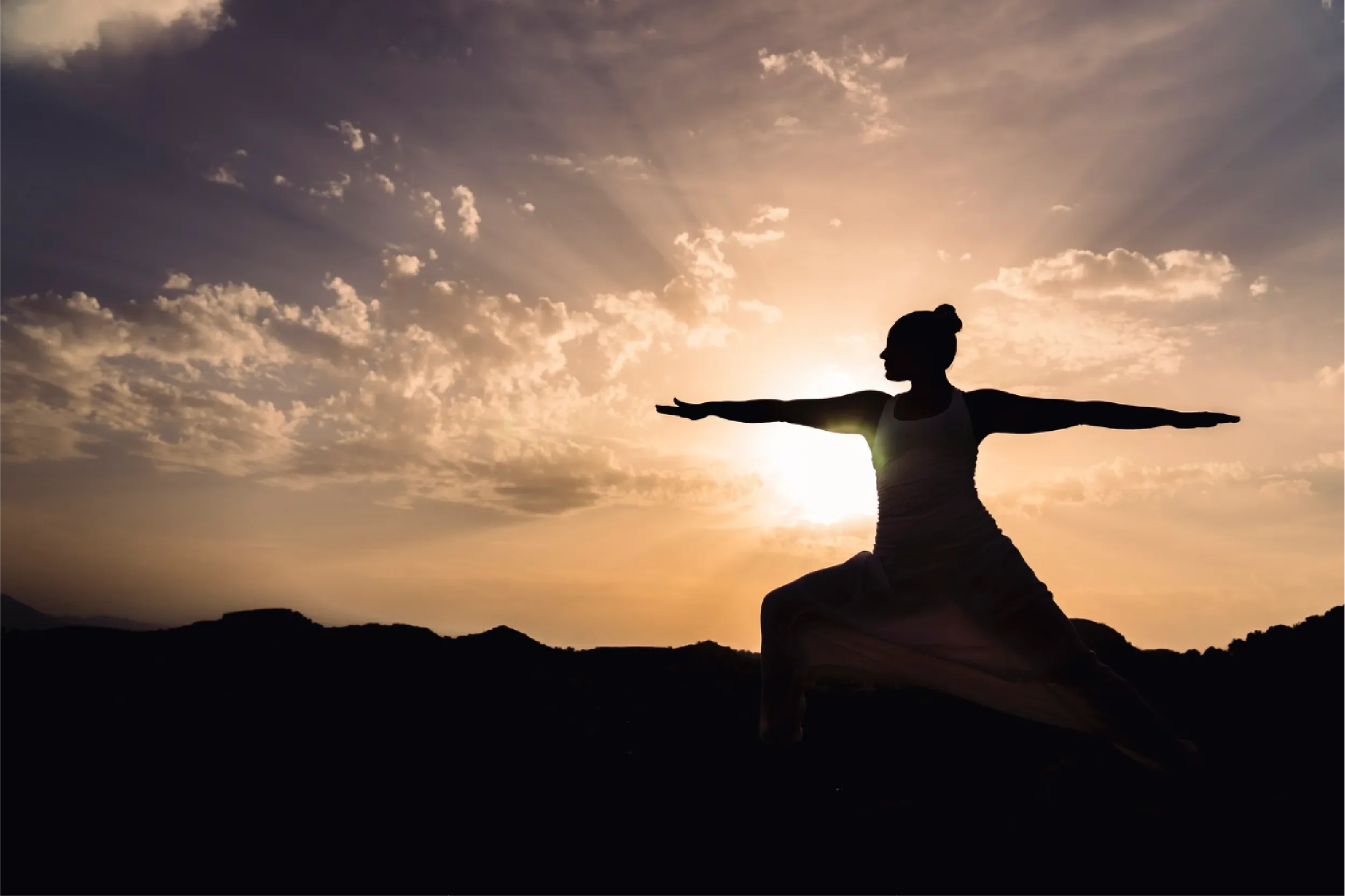 Silhouette of a woman practicing a yoga warrior pose against a dramatic sunset sky with radiant sun rays, representing Universal Human Foundation's support for spiritual growth, balance, and inner strength.