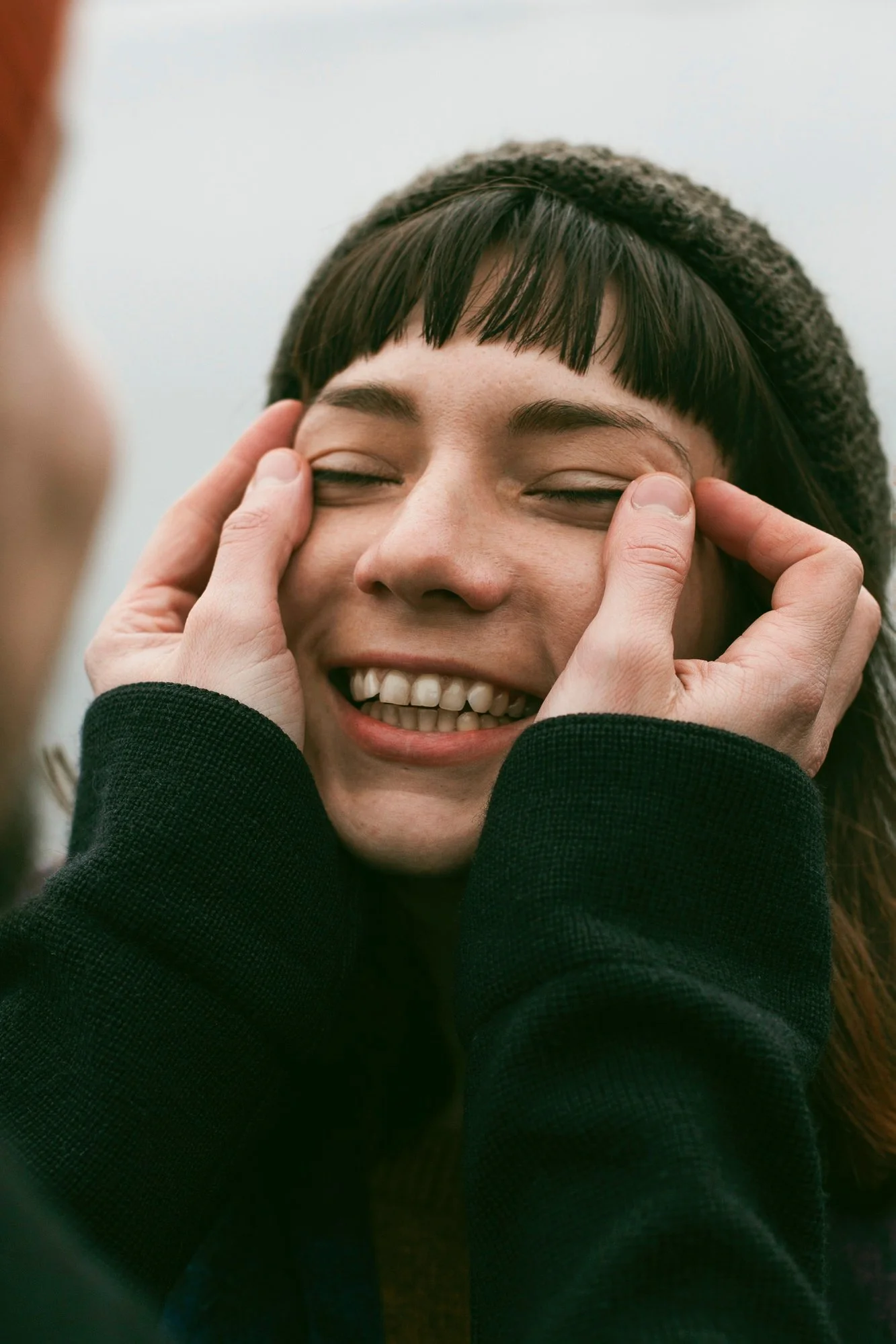 Young woman with a beanie laughing with pure joy, hands framing her face, representing Universal Human Foundation's support for authentic joy, emotional freedom, and self-expression.