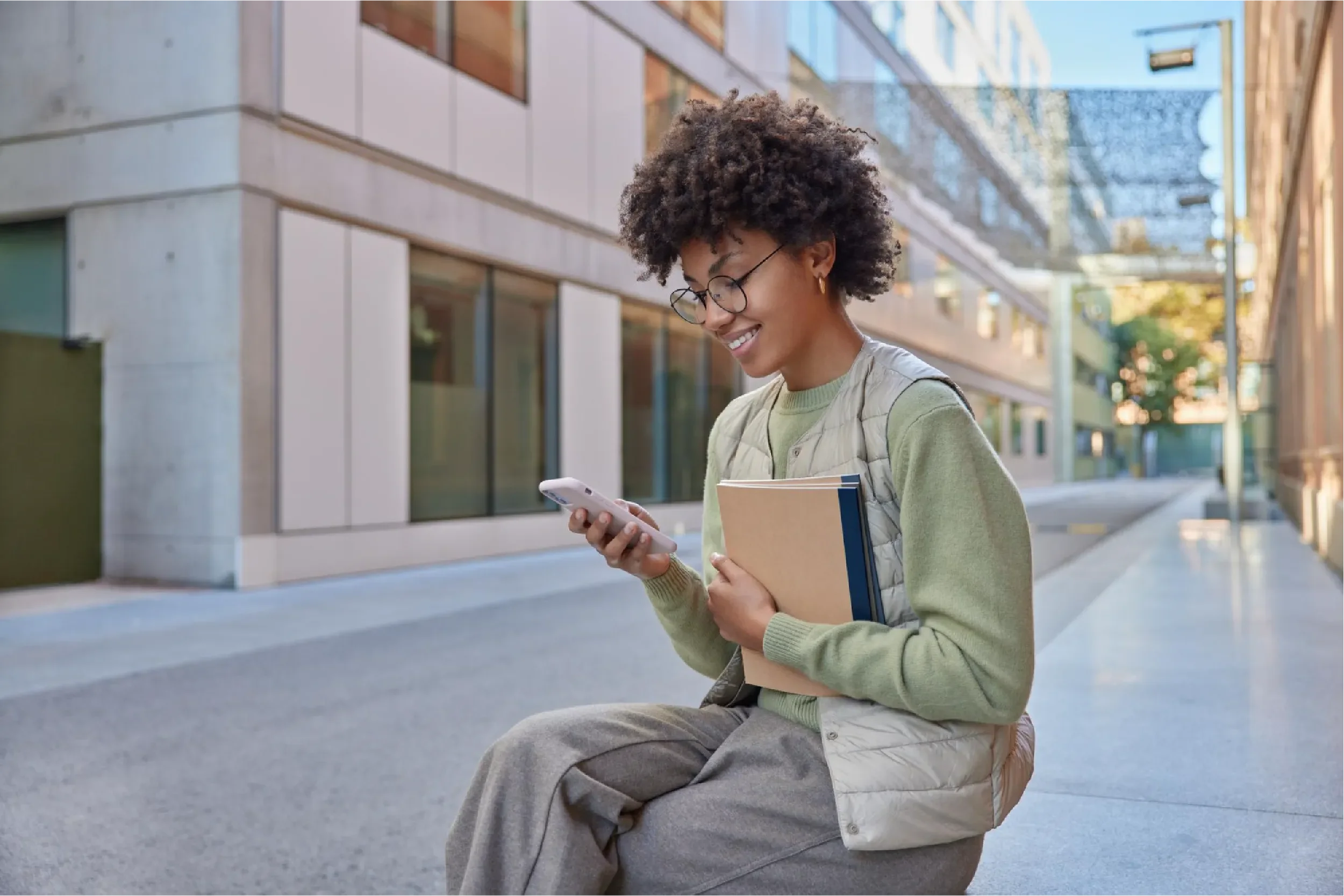 Young student with curly hair and glasses smiling while looking at her smartphone and holding books in an urban setting, representing Universal Human Foundation's support for students and young adults navigating modern life.