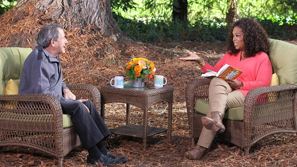 Gary Zukav and Oprah Winfrey seated outdoors in a garden setting, engaged in a warm conversation, with one holding an open book, surrounded by trees and a small table with flowers, reflecting connection, reflection, and meaningful dialogue