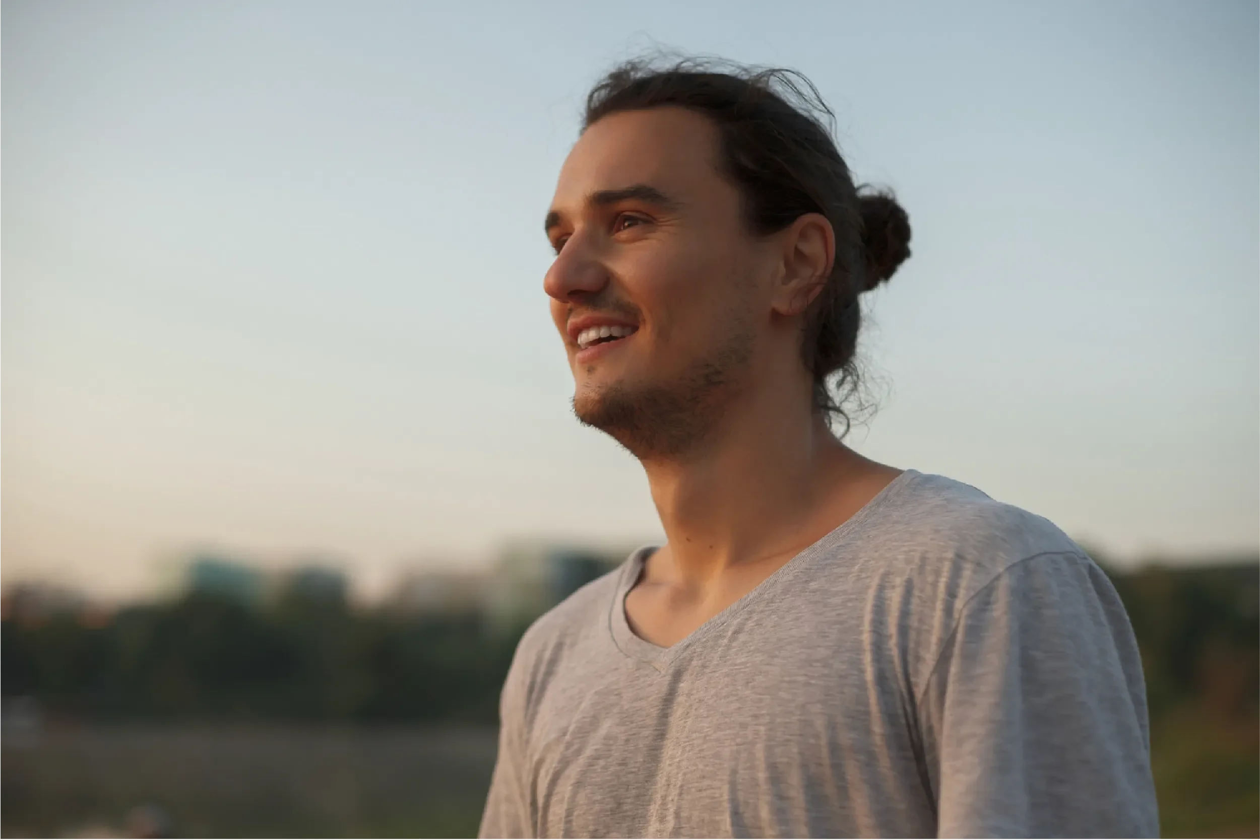 Young man with a man bun smiling contentedly outdoors at golden hour, representing Universal Human Foundation's support for authentic self-expression, joy, and personal growth.