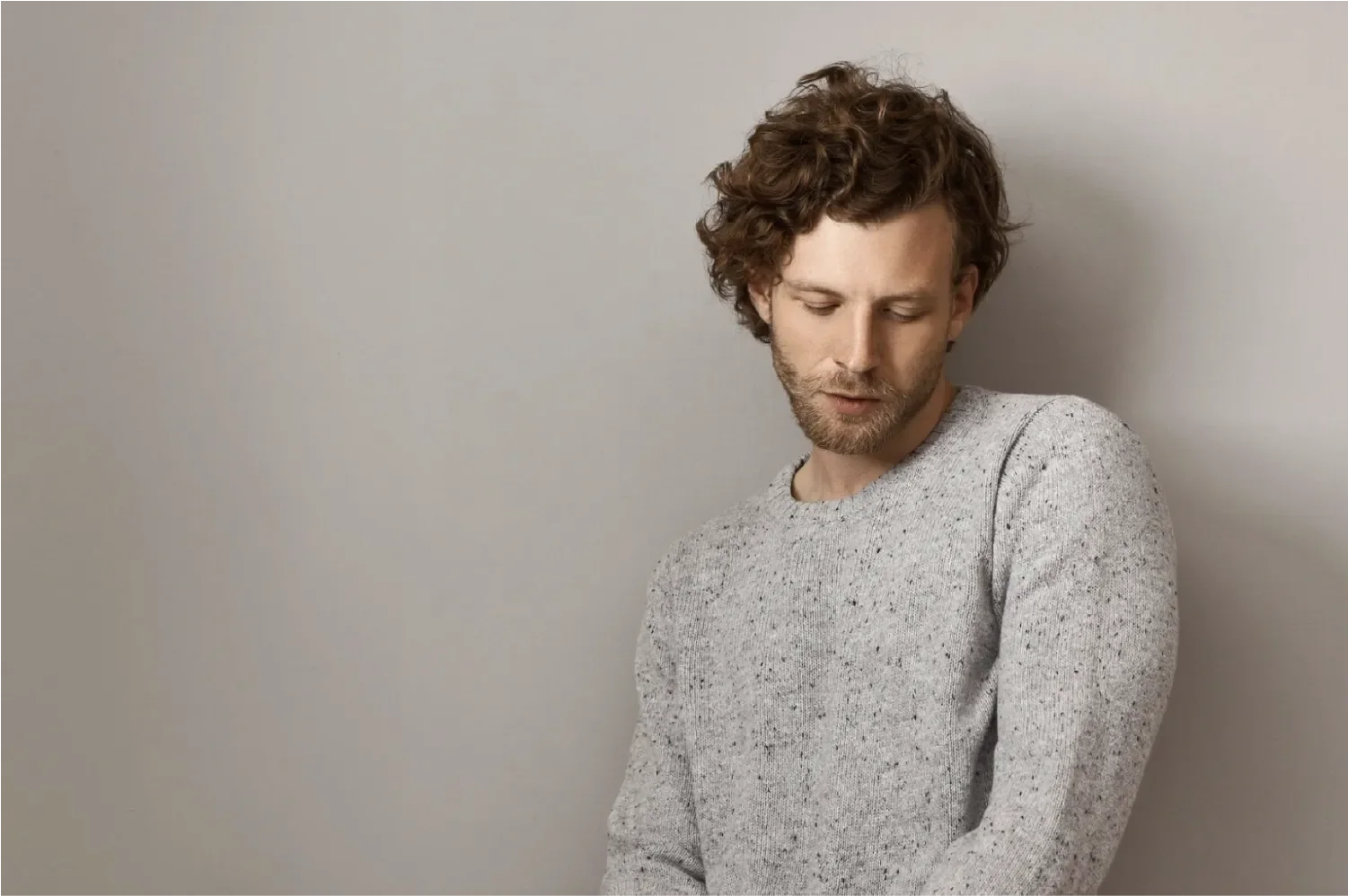 Young man with curly hair in a grey sweater looking down thoughtfully against a neutral background, representing Universal Human Foundation's support for introspection, emotional awareness, and personal growth.
