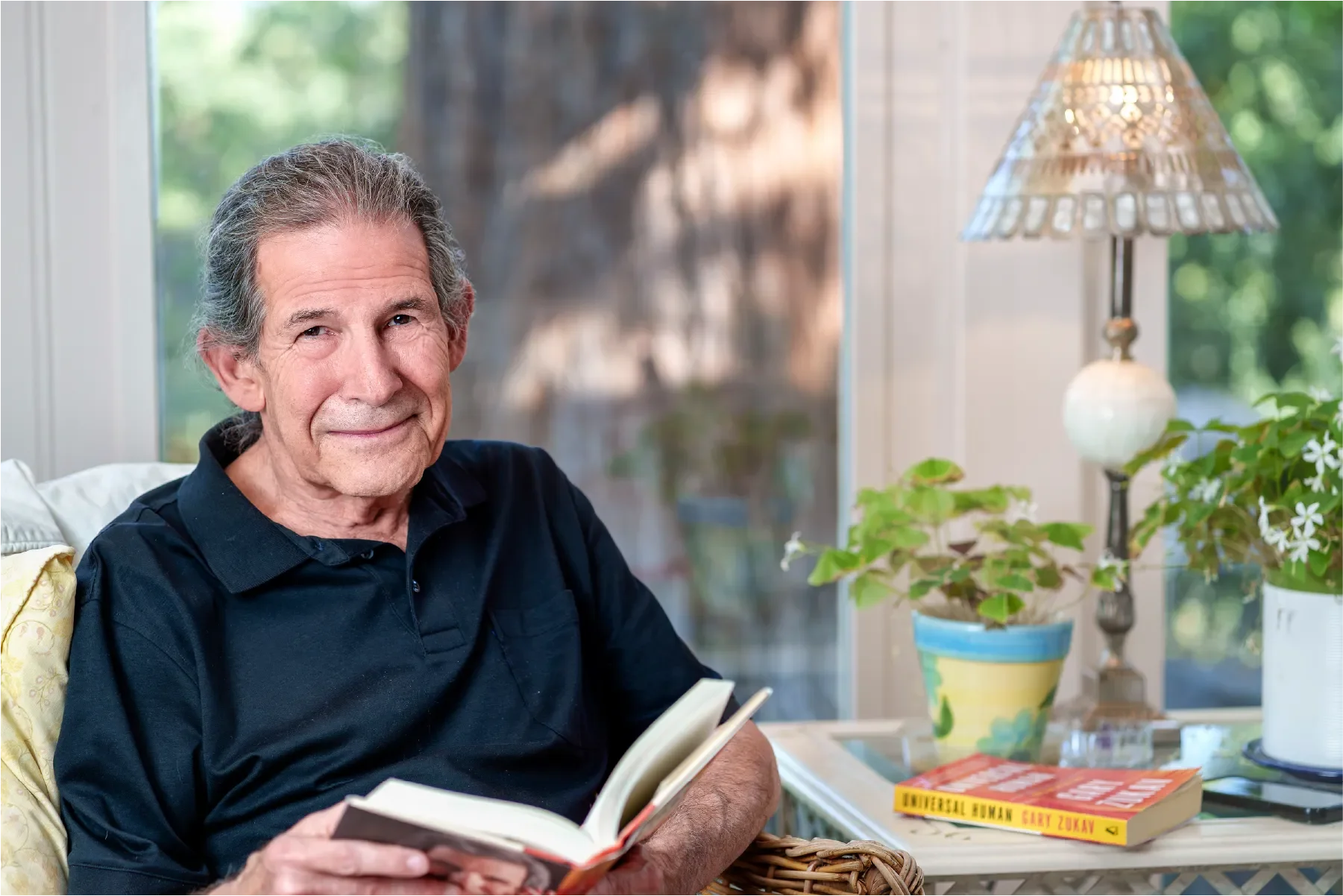 ary Zukav, author and co-founder of Seat of the Soul Institute, smiling while reading a book at home, with his book "Universal Human" visible on the side table.