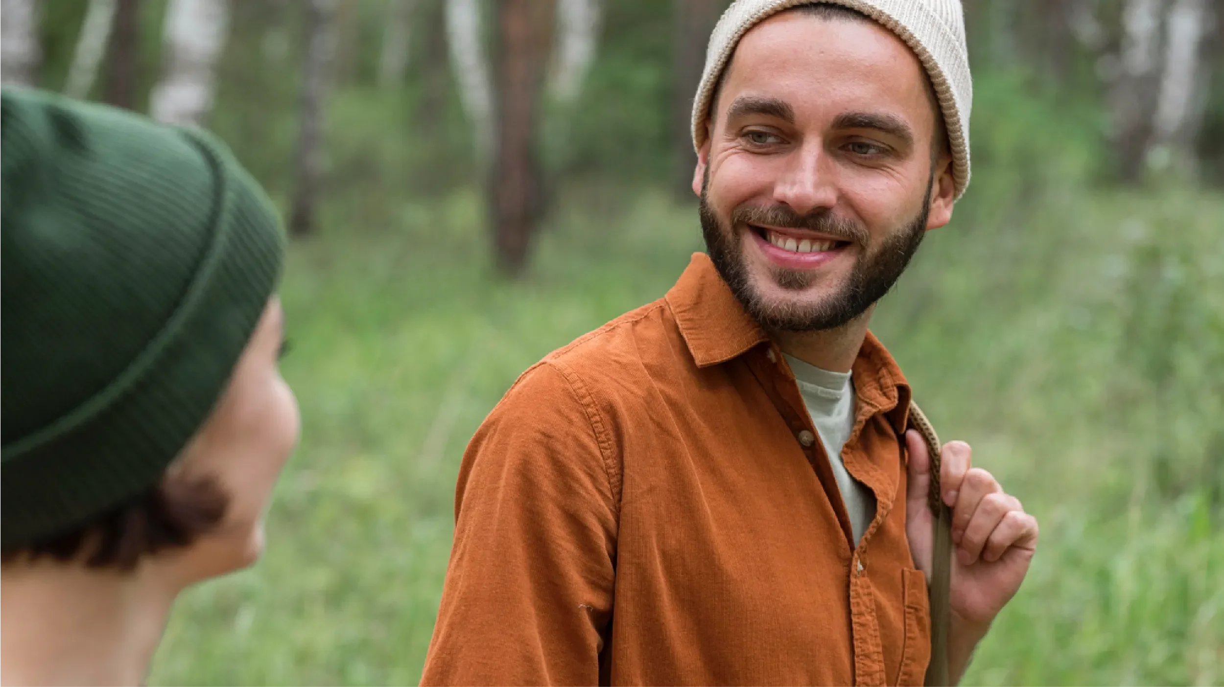 Young man in a beanie and orange shirt smiling warmly during a conversation with a friend in a forest setting, representing Universal Human Foundation's support for authentic connection and meaningful relationships.