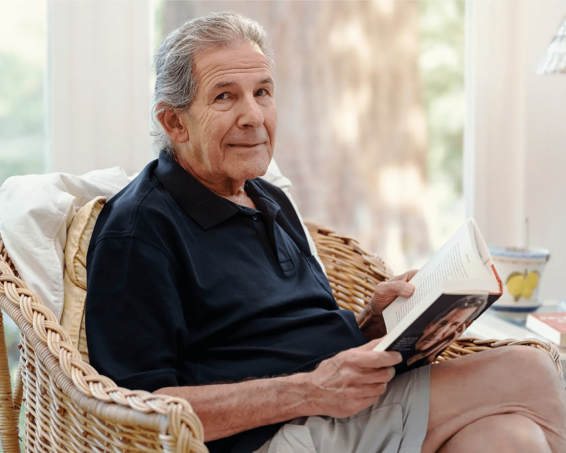 Gary Zukav, author and co-founder of Seat of the Soul Institute, sitting in a wicker chair holding an open book, looking thoughtfully into the distance.