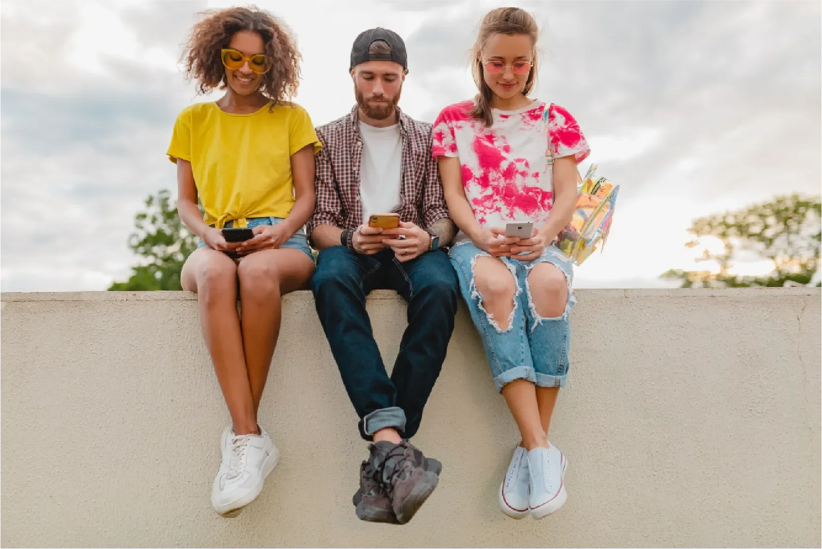 Three young millennial adults sitting on a wall outdoors, each looking at their smartphones, representing Universal Human Foundation's support for navigating economic pressure, identity, and connection in a fast-changing world.