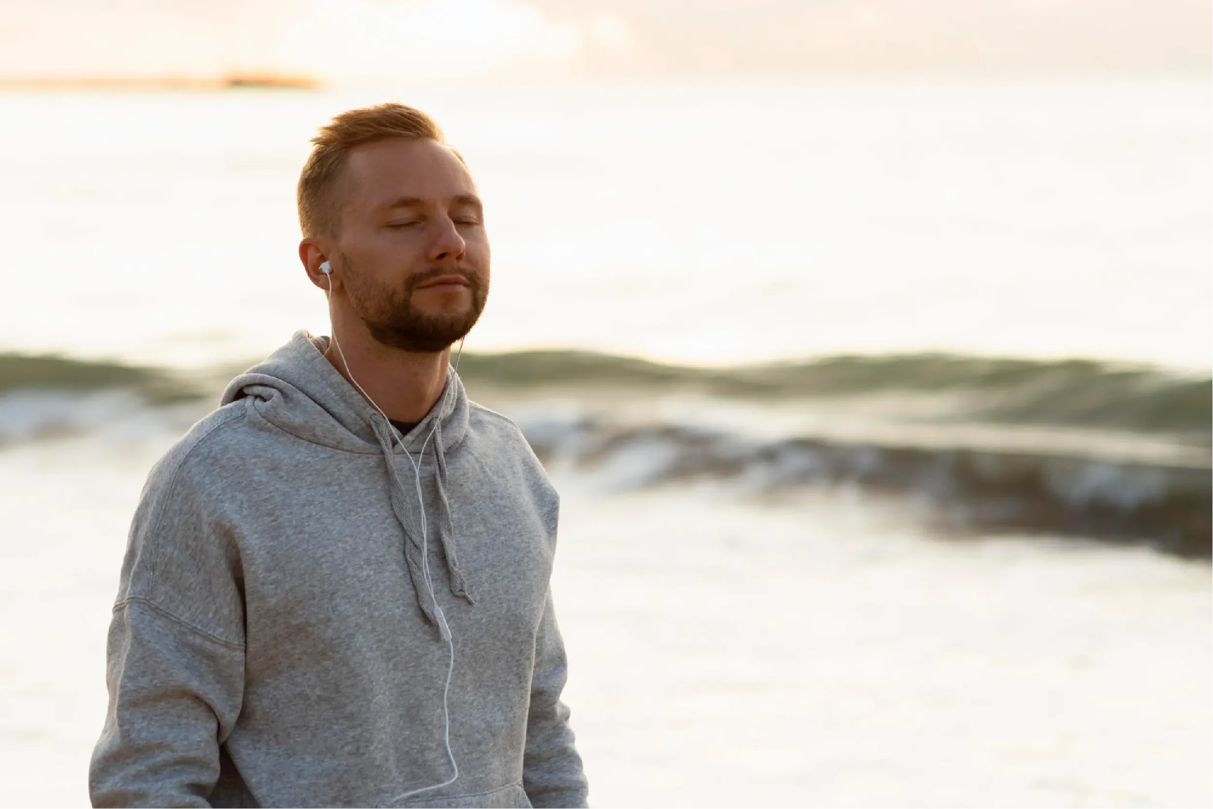 Young man in a grey hoodie with earphones meditating peacefully by the ocean at sunset, representing Universal Human Foundation's support for mindfulness, inner calm, and emotional wellbeing.