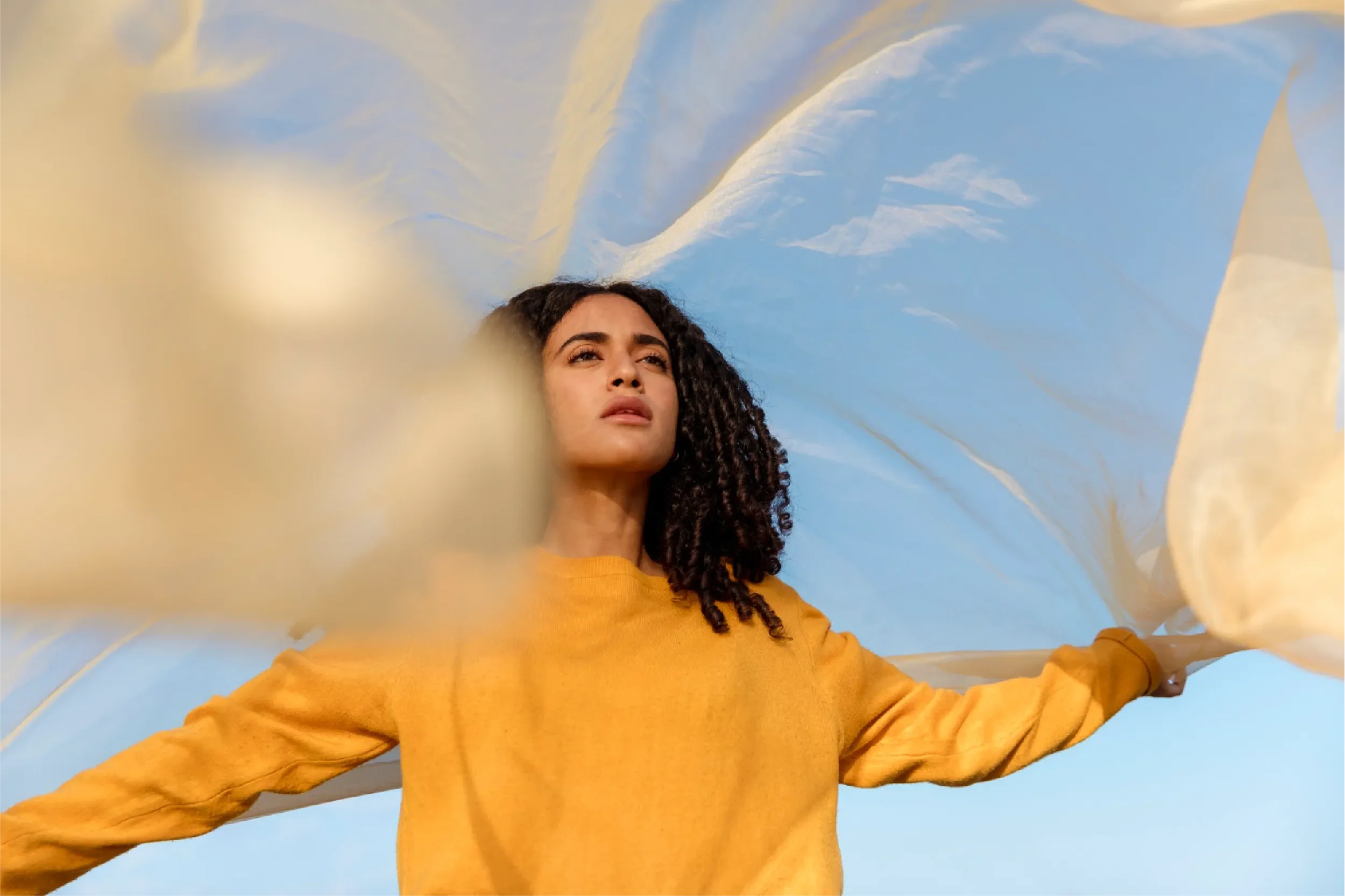 Young woman in a yellow sweater with curly hair standing confidently with arms outstretched, holding flowing fabric against a bright blue sky, representing Universal Human Foundation's support for freedom, empowerment, and authentic living.