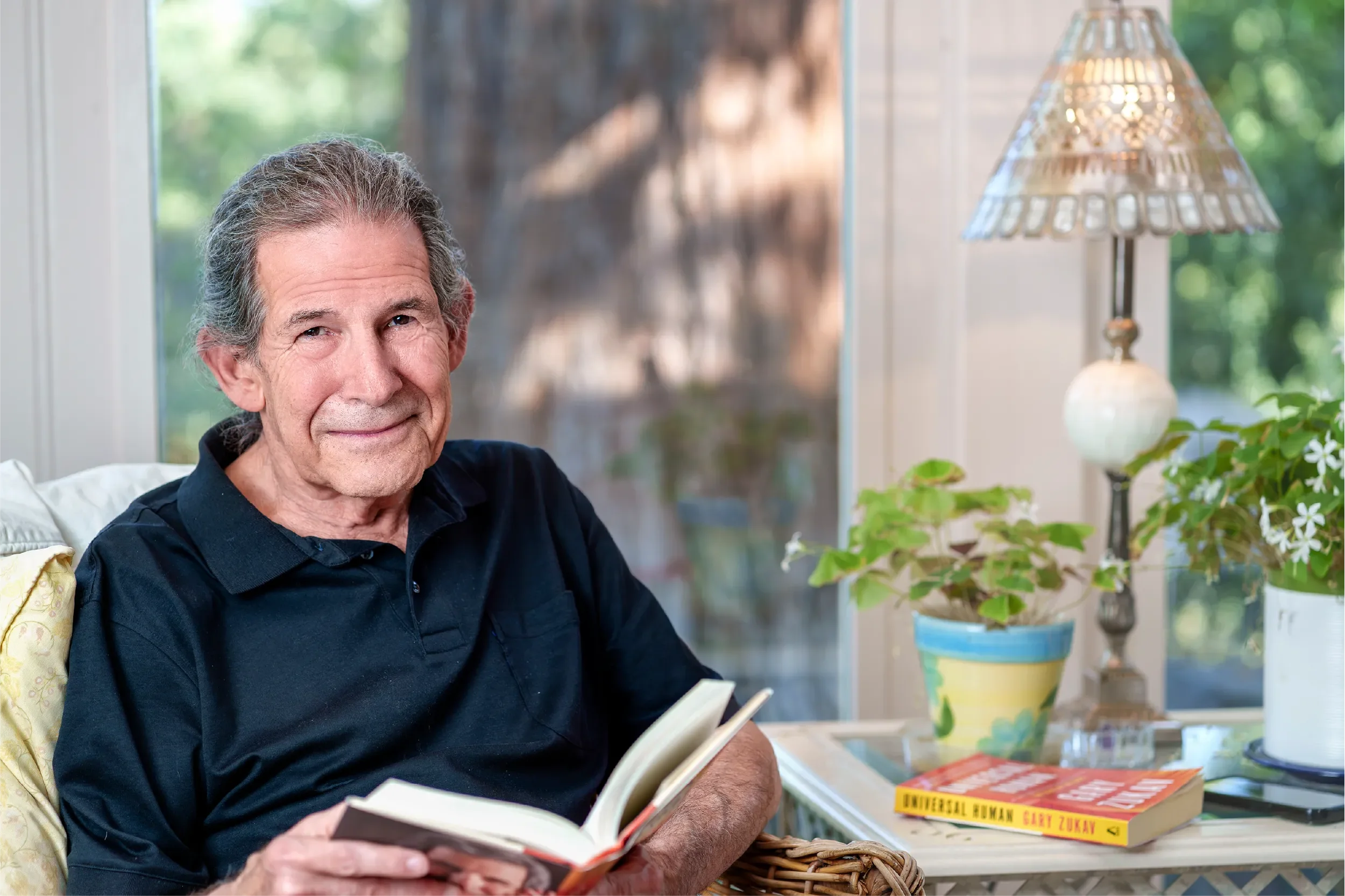 Gary Zukav, author and co-founder of Seat of the Soul Institute, smiling warmly while reading a book at home, with his book "Universal Human" visible on the side table.
