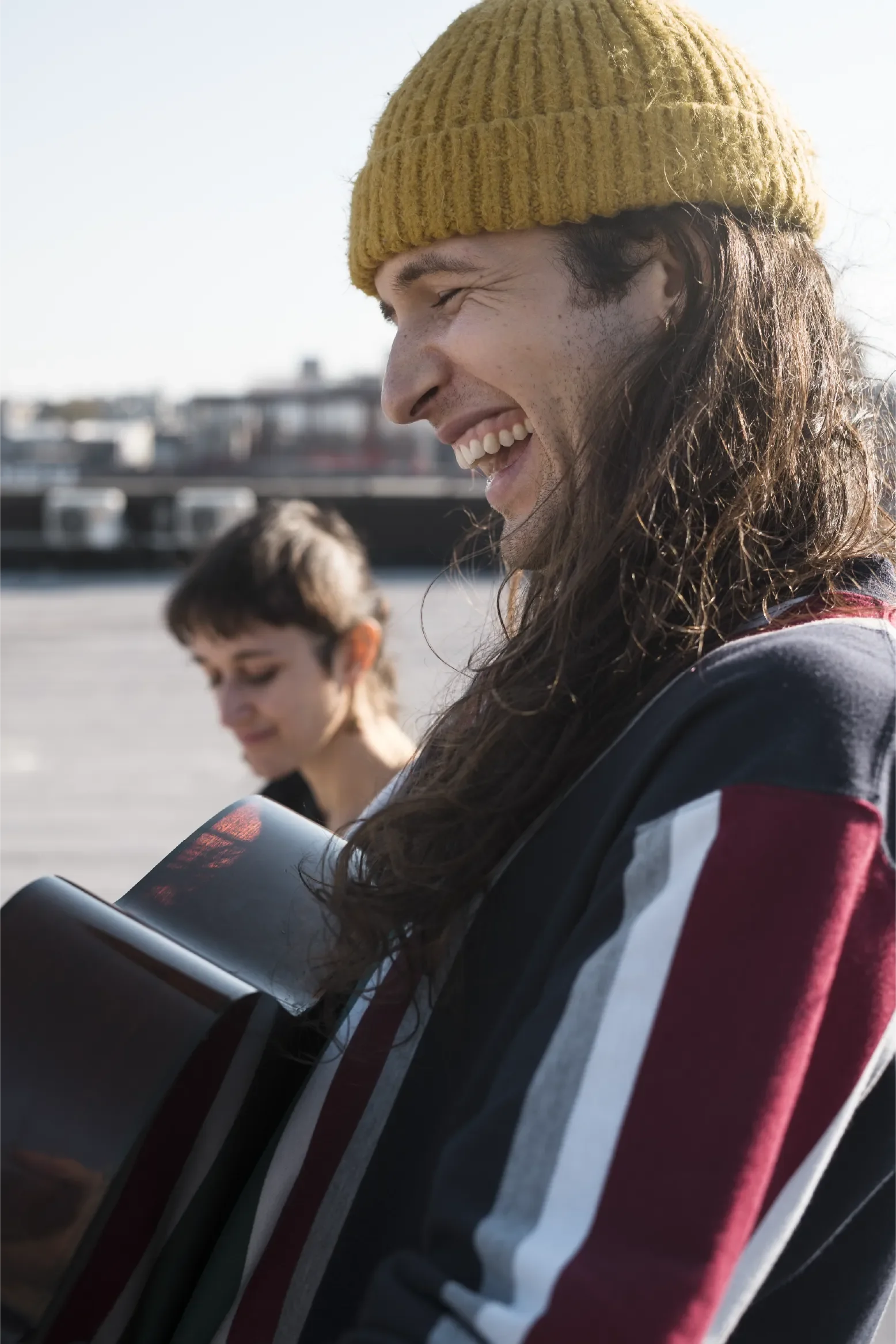Smiling person with long hair wearing a knit beanie, holding a guitar outdoors near the water, with another person in the background, capturing a moment of connection, creativity, and shared presence