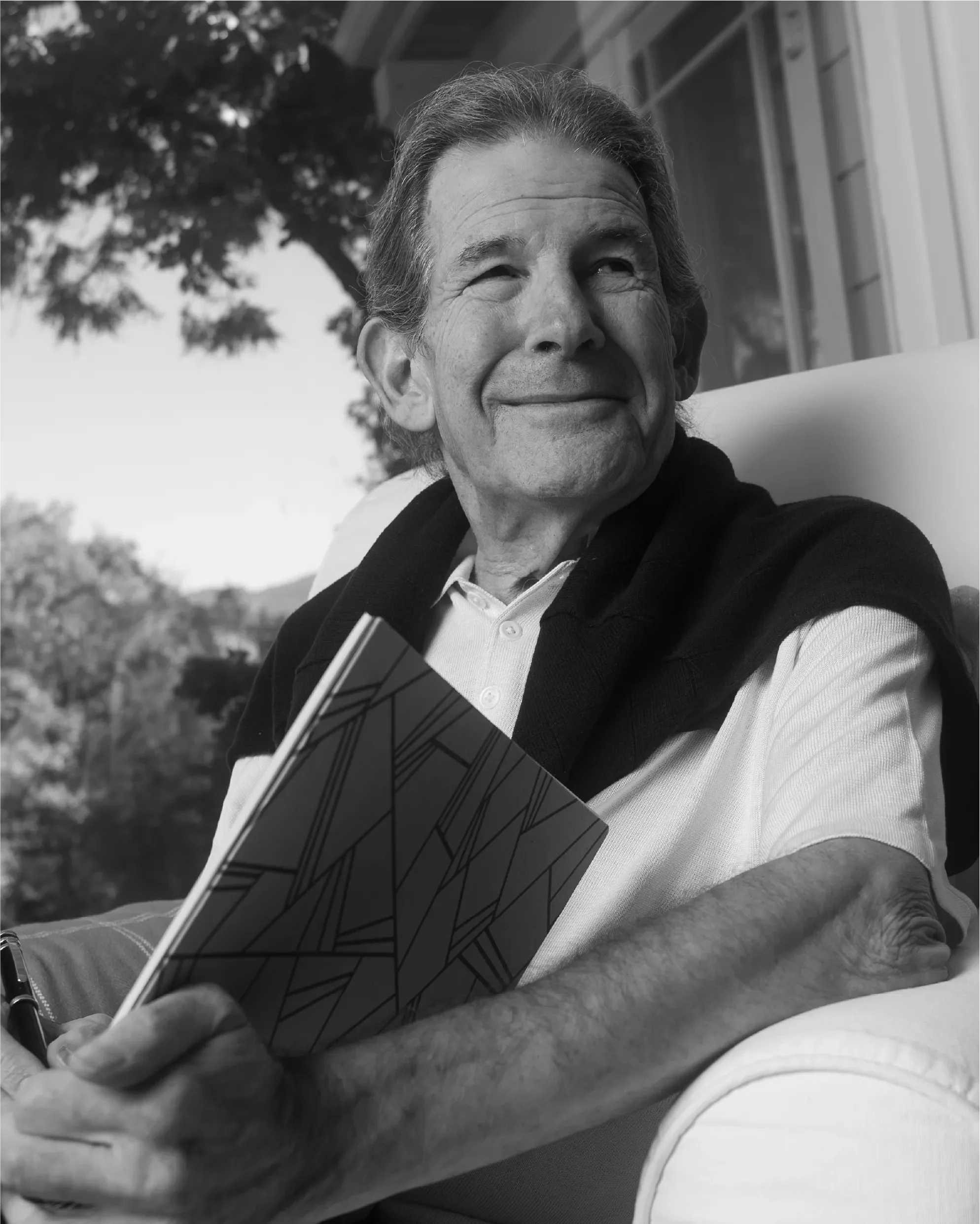 Black-and-white portrait of Gary Zukav, author and co-founder of Seat of the Soul Institute, smiling gently while holding a notebook outdoors.