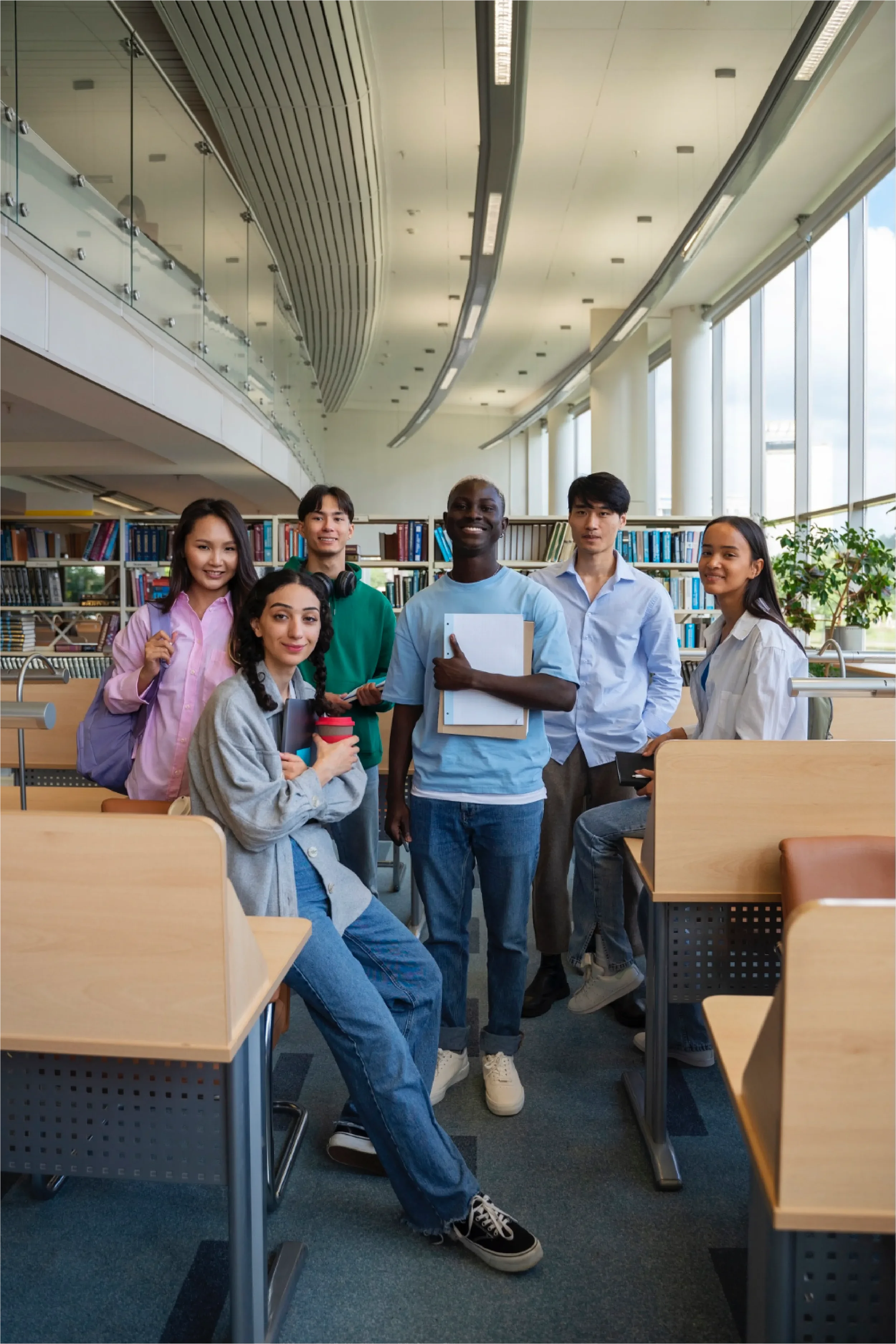 Diverse group of six university students standing together in a modern library with bookshelves in the background, representing Universal Human Foundation's support for students, education, and meaningful community.