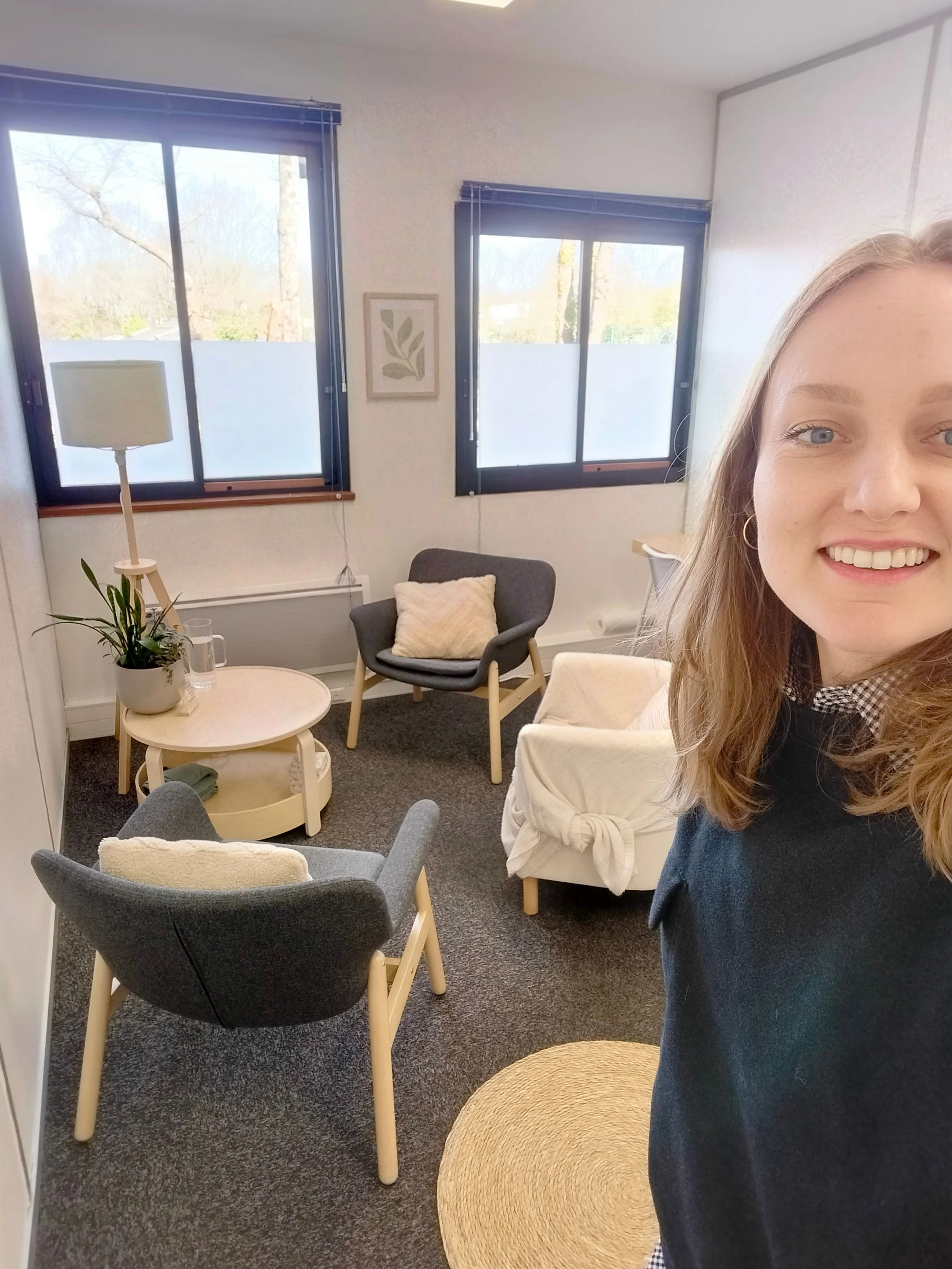 Une jeune femme souriante dans un salon confortable avec plusieurs fauteuils, une table ronde, une lampe, et deux fenêtres laissant entrer la lumière naturelle