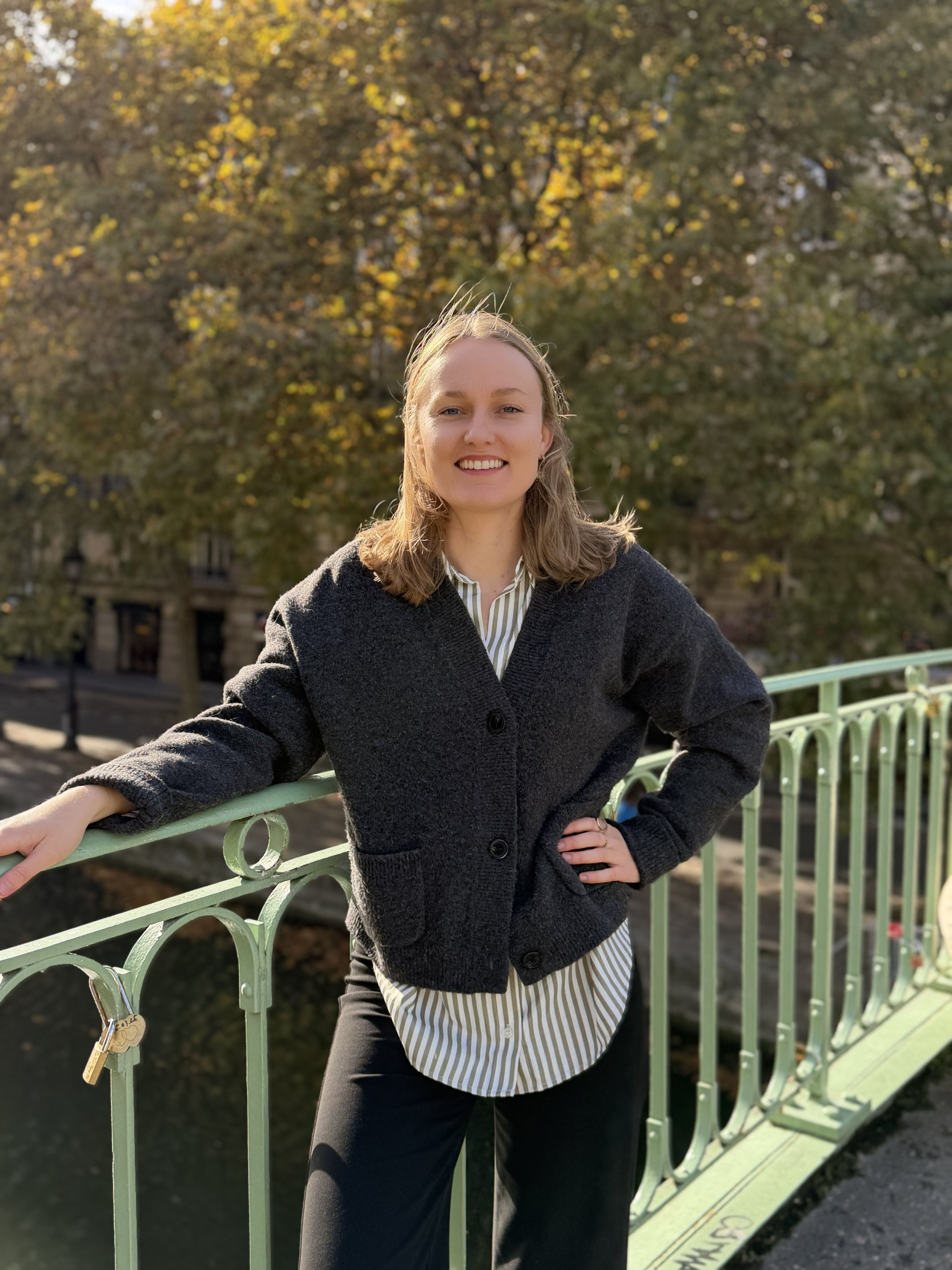 Jeune femme souriante posant sur un pont en fer vert avec des arbres aux feuilles automnales en arrière-plan.