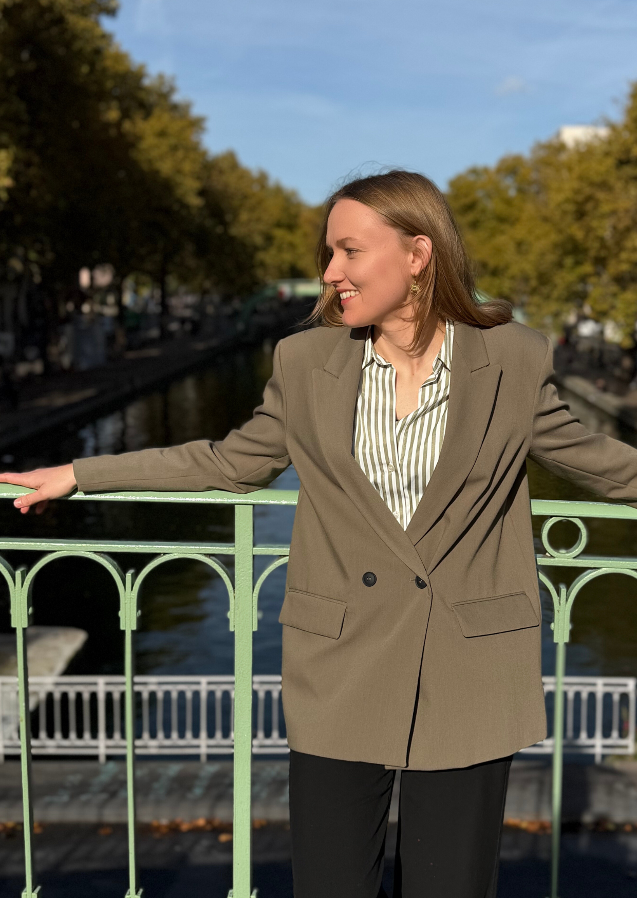 Une femme souriante portant un blazer beige et une chemise à rayures, se tient sur un pont à côté d'une rivière, avec des arbres et un ciel bleu en arrière-plan.