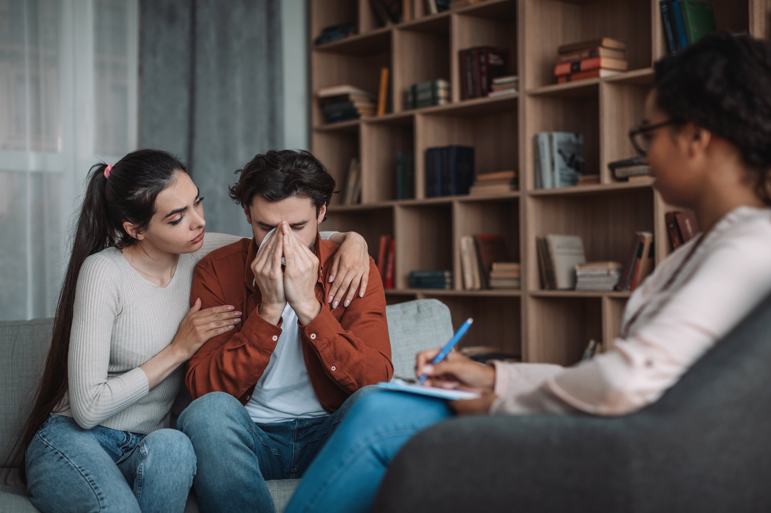 A woman and a man on a couch, with the man holding his head and a tissue to his face, appearing distressed. Another woman is sitting across from them, taking notes, in a room with bookshelves in the background.