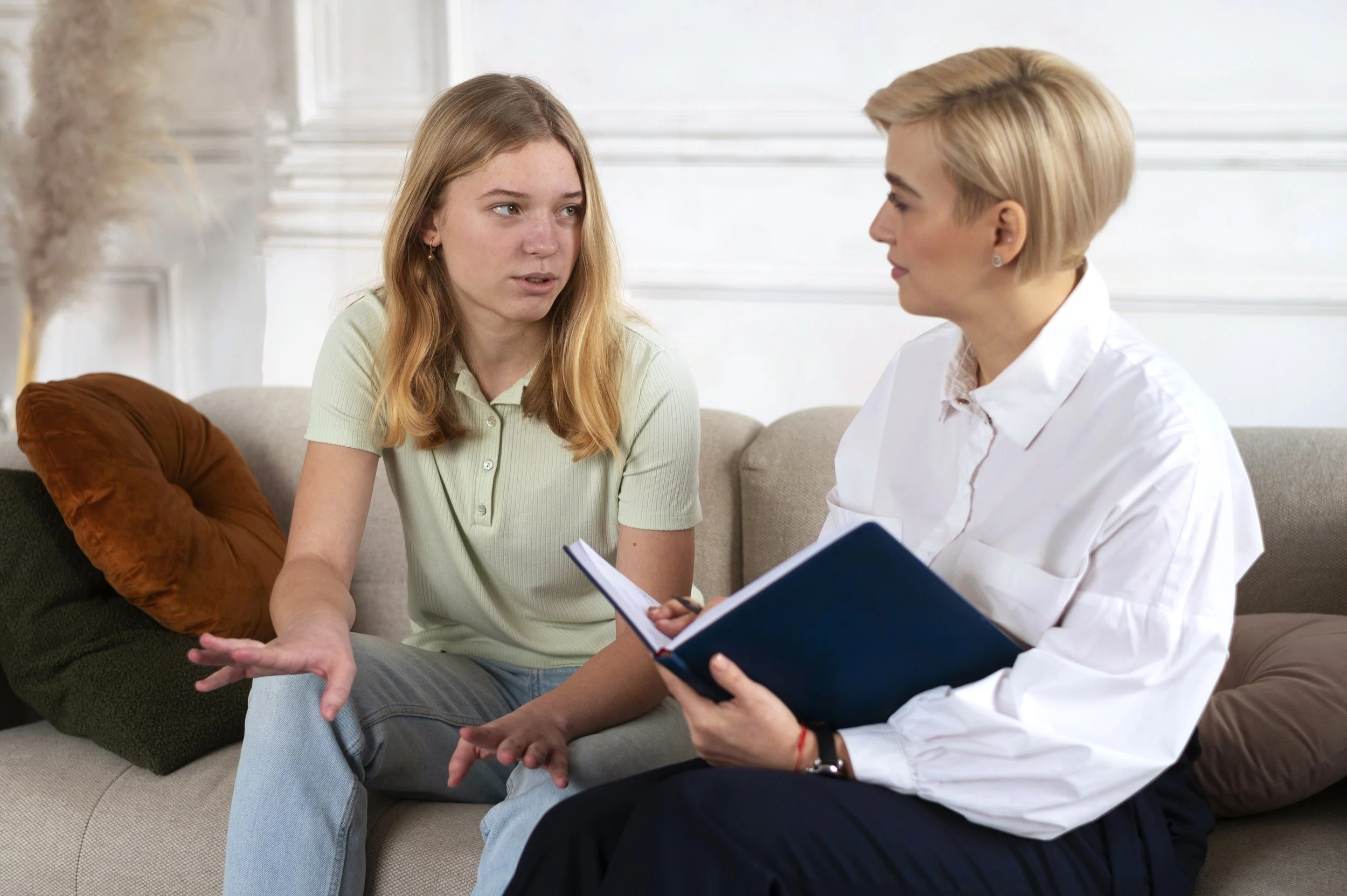 Young woman talking to an older woman during a counseling session, sitting on a beige couch with pillows, the older woman holding a notebook.