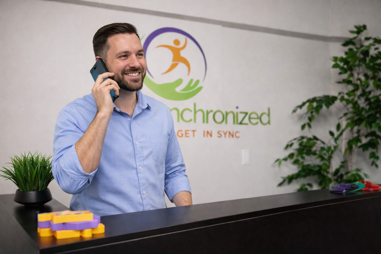 A smiling man with dark hair and a beard, wearing a light blue shirt, talking on a cell phone behind a reception desk. There is a small potted plant and colorful tokens on the desk. Behind him, a logo with a stylized figure and the words "Chronized" and "GET IN SYNC" are on the wall.