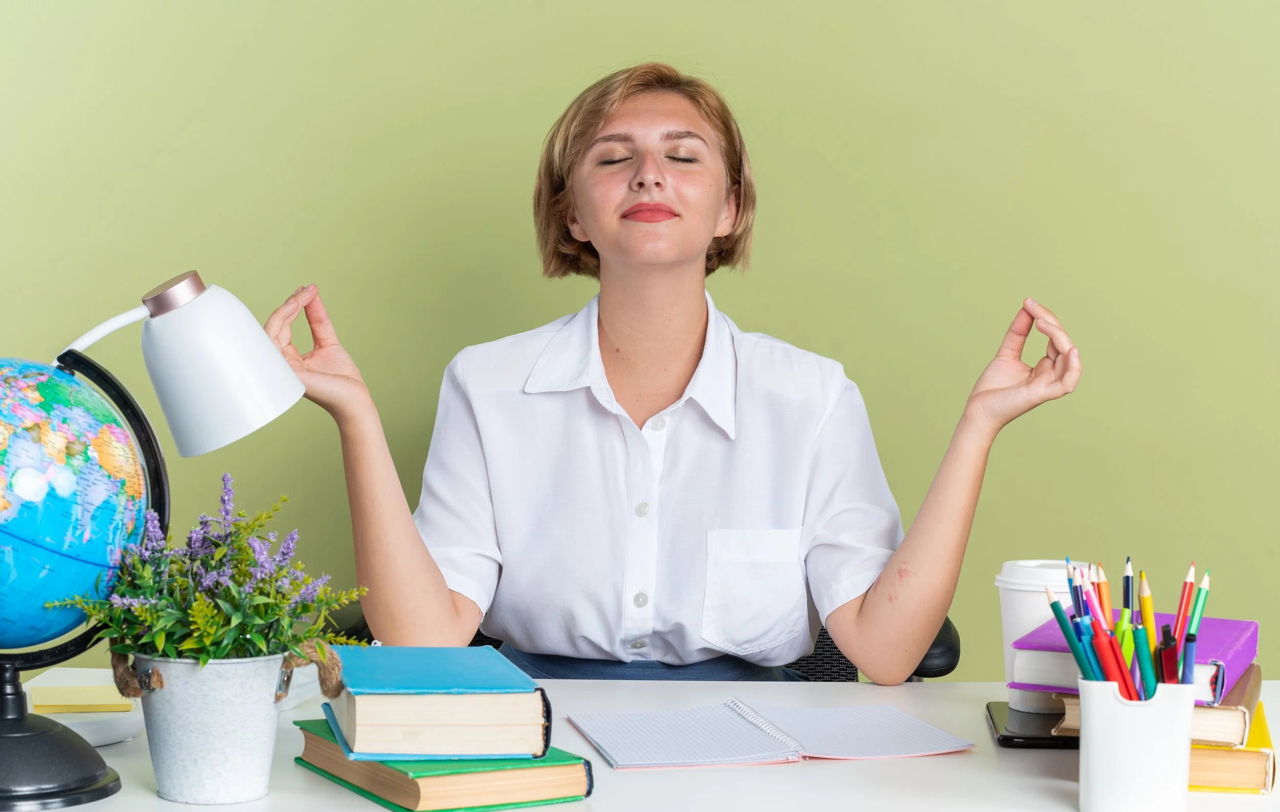 Woman practicing meditation at a desk with books, globe, plants, and stationery.