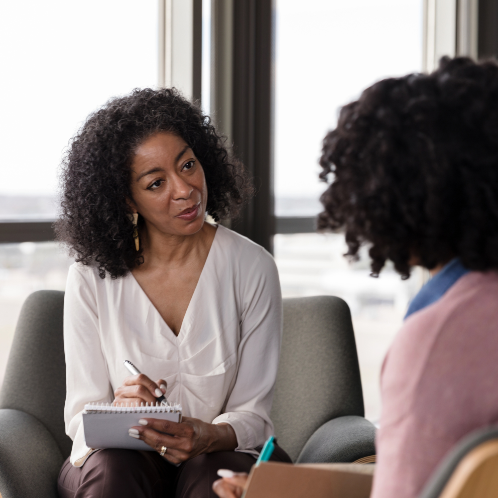 Two women having a conversation indoors, one taking notes with a notepad, in a professional setting with large windows in the background.