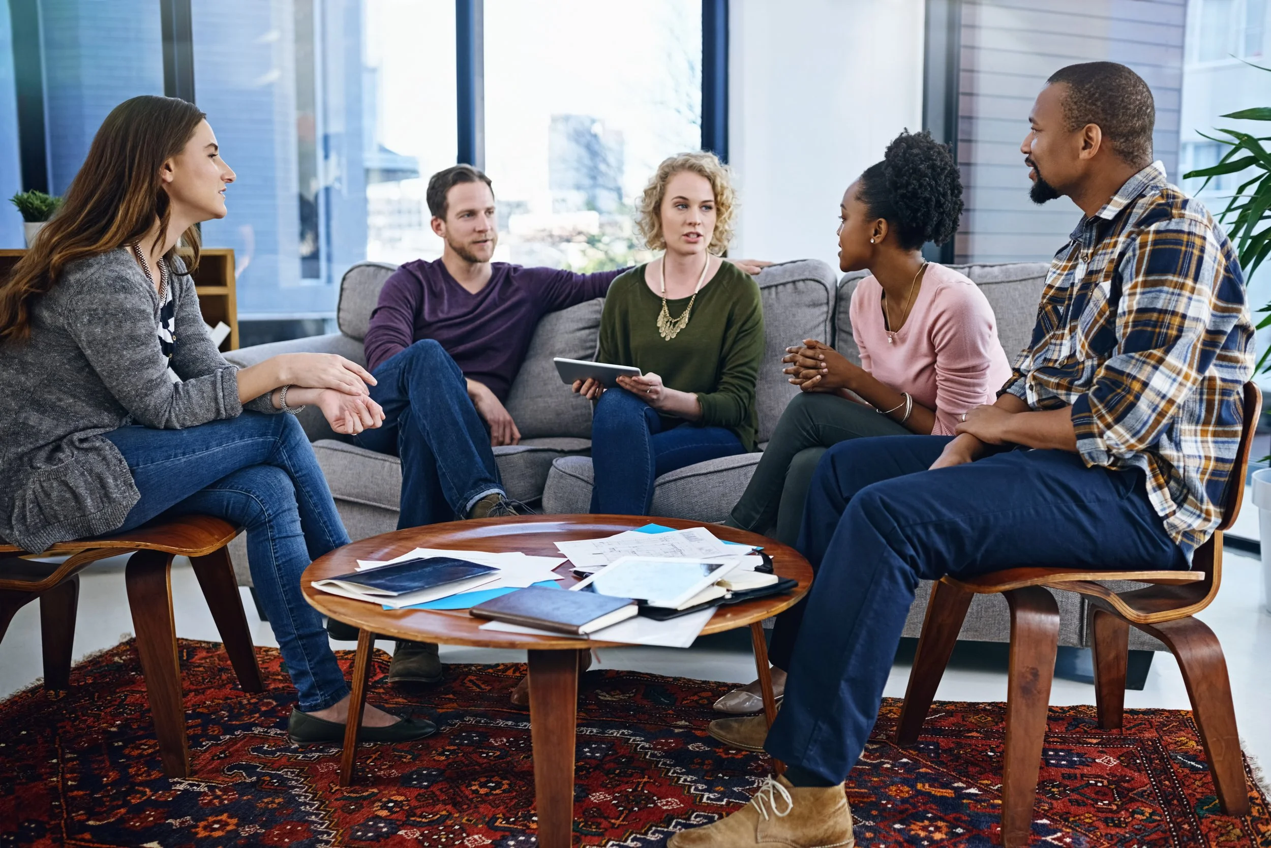 Six diverse people having a discussion in a bright, modern living room with large windows, sitting on a sofa and chairs around a wooden coffee table with papers and tablets on it.