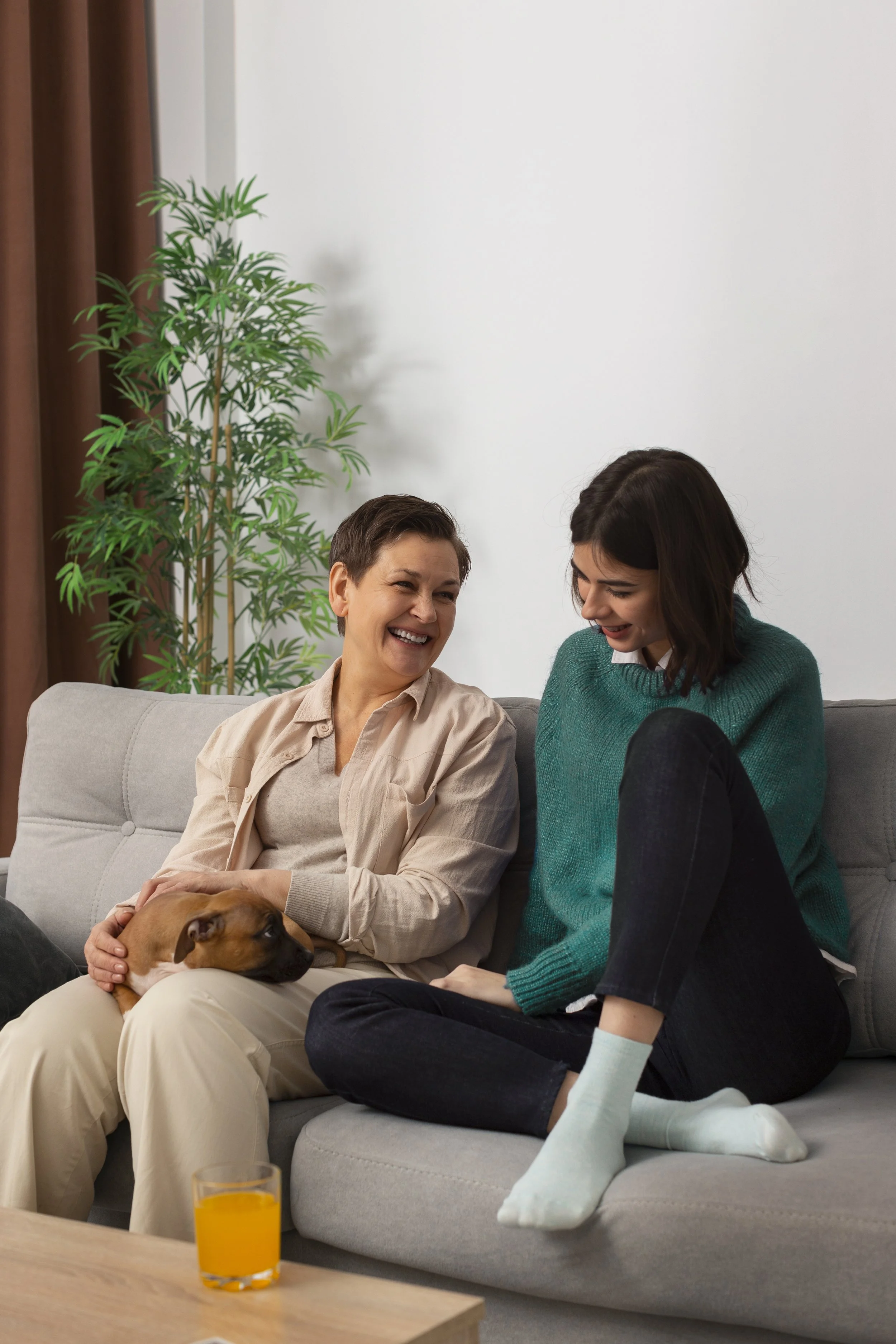 Two women sitting on a gray sofa, smiling and laughing, with a brown dog on the woman's lap. A glass of orange juice is on the table in front of them, and a green plant is in the background.