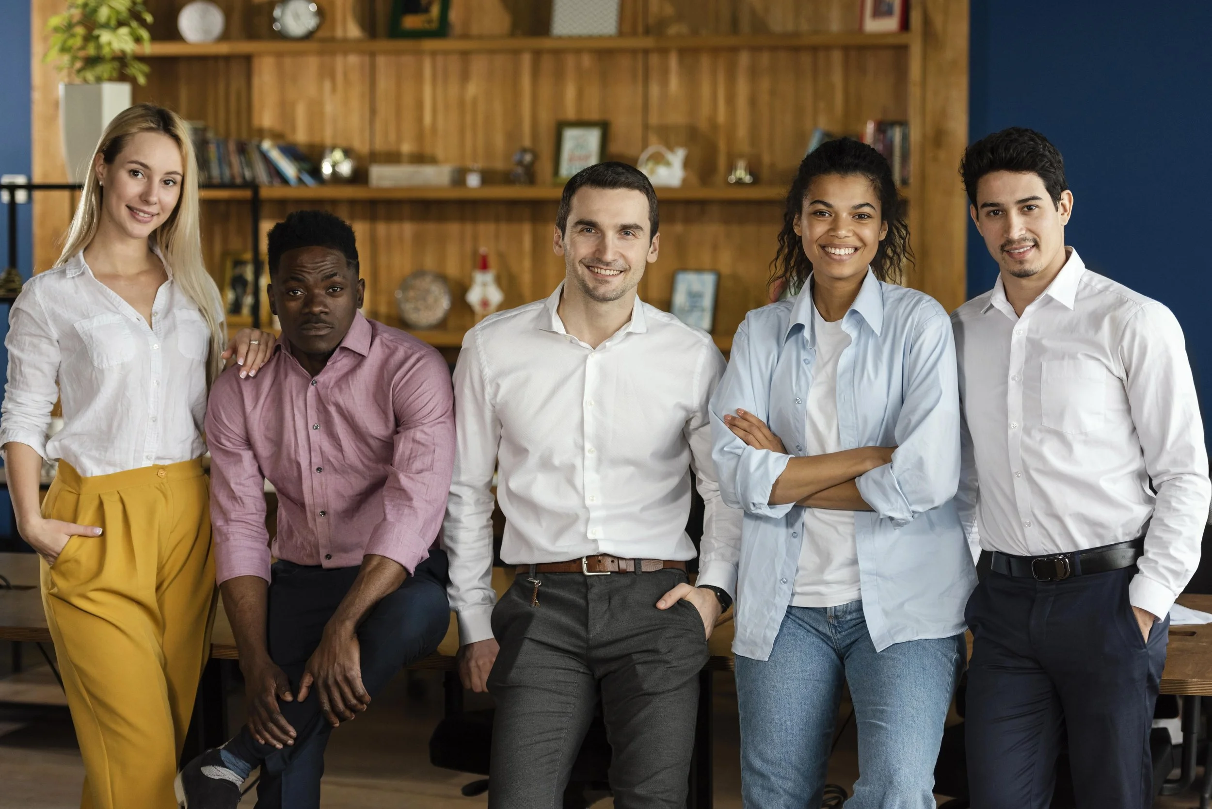 Group of five diverse young professionals standing together and smiling in an office setting with wooden shelves and decorations behind them.