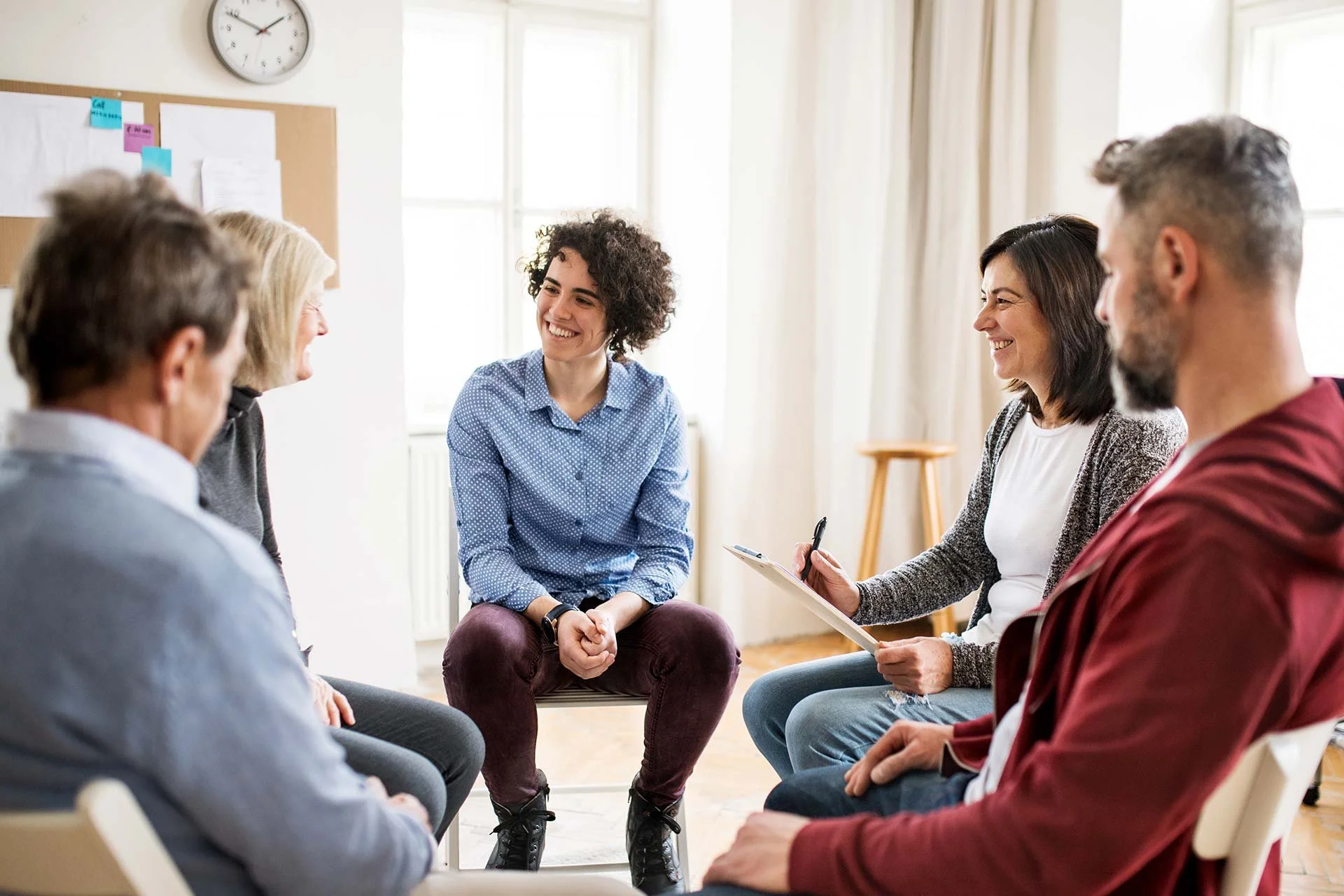 Five people sitting in a circle having a conversation in a bright room, smiling.