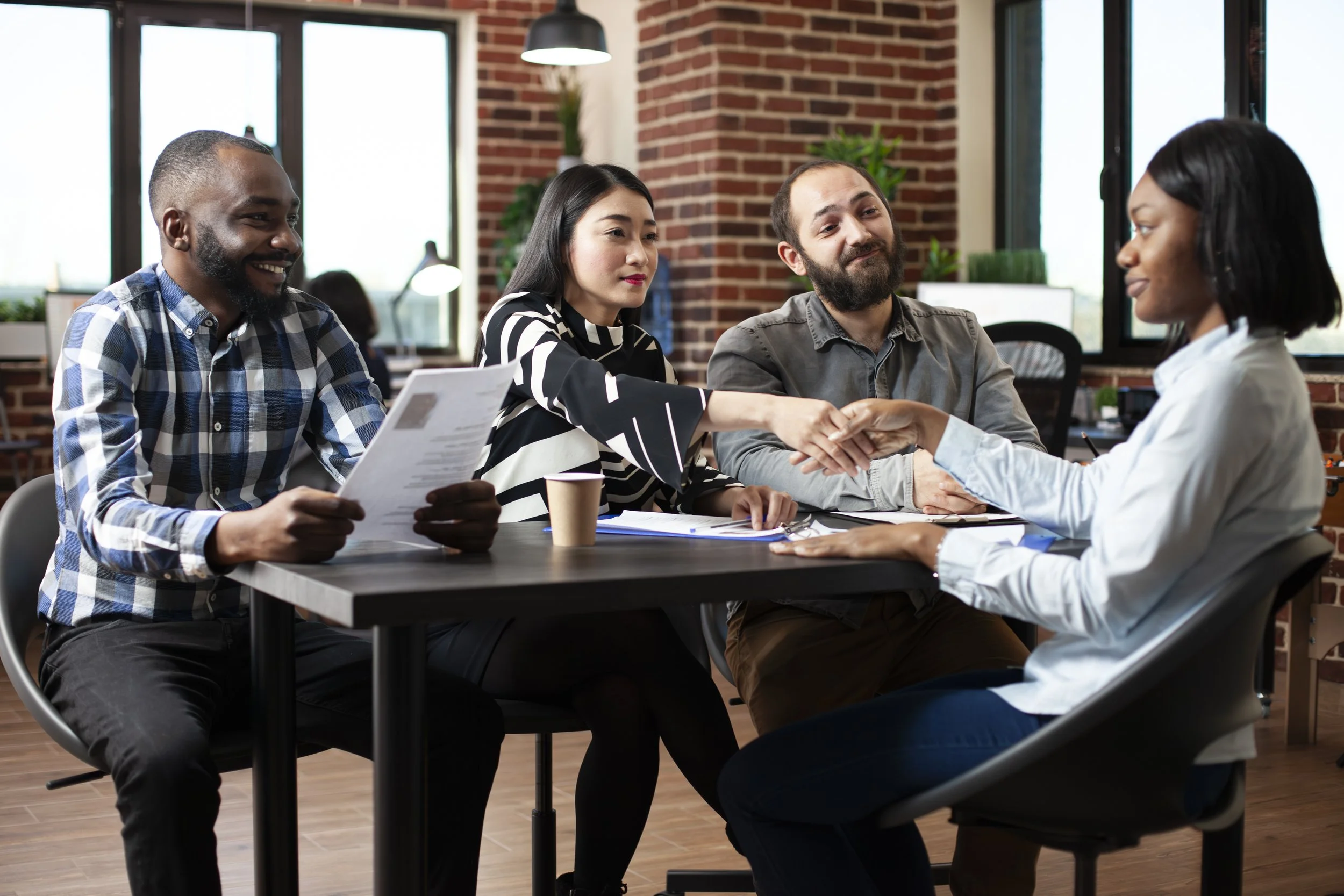 Four diverse people in a professional setting, two men and two women, engaging in a business meeting with one woman shaking hands with another.