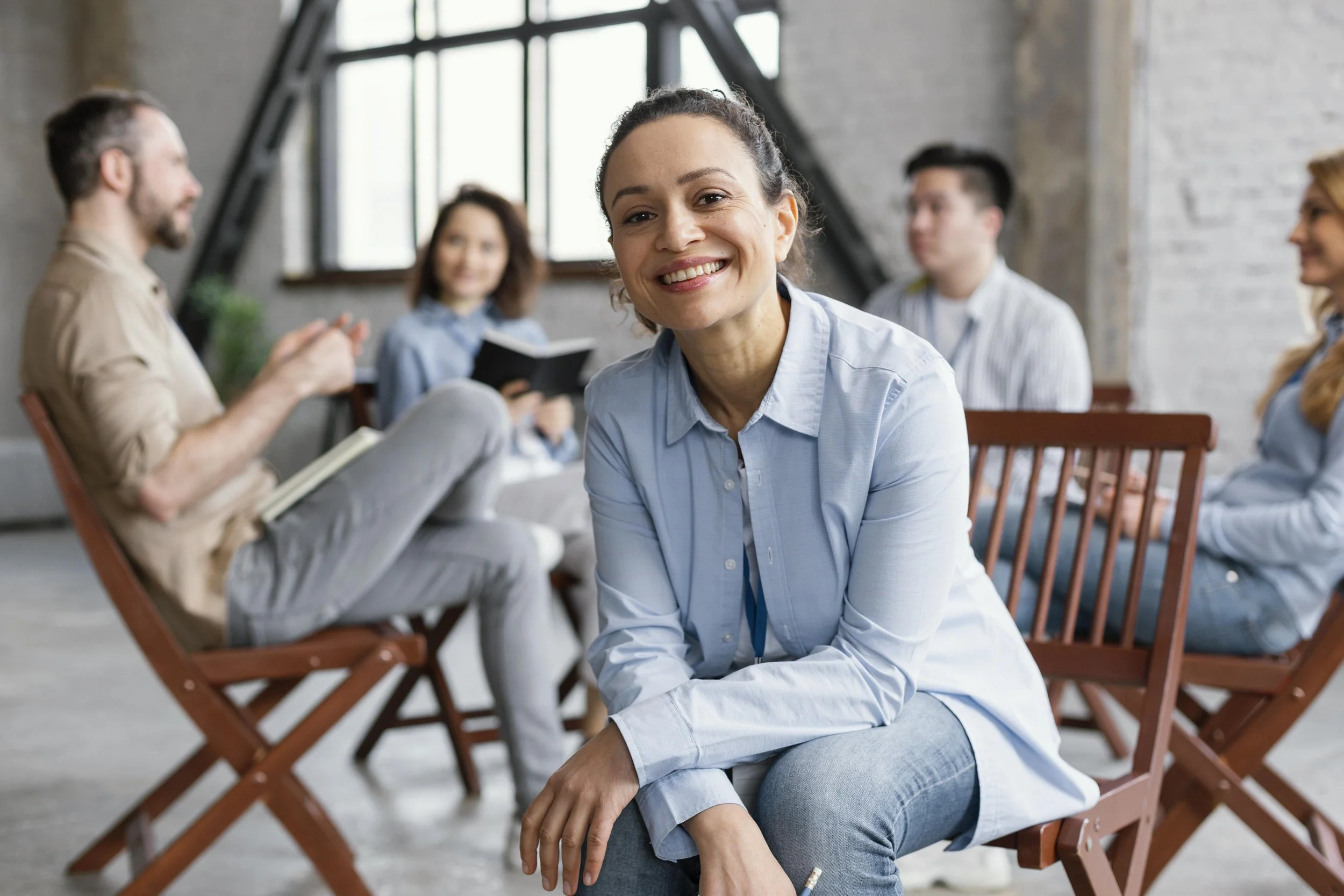 A woman is smiling and sitting on a wooden chair in a meeting or seminar setting, with other people sitting and engaged in discussion in the background.