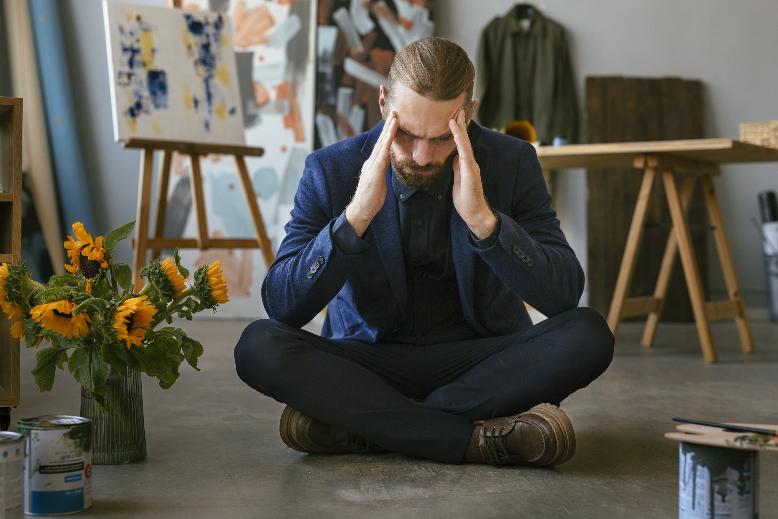 A man sitting cross-legged on the floor, holding his head with both hands, appearing stressed or anxious, in an art studio with paintings and art supplies around.