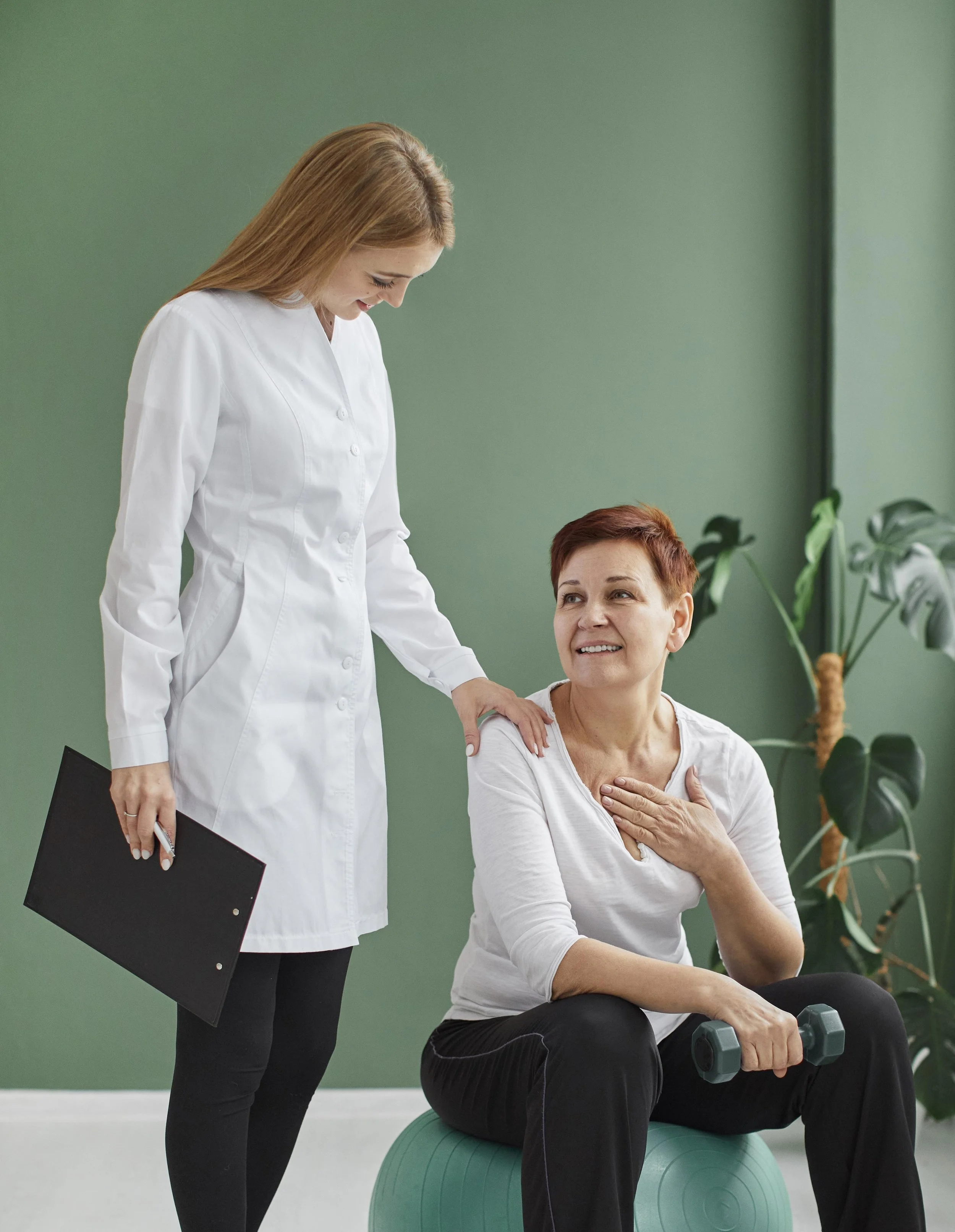 A female physical therapist talking to a woman exercising with dumbbells on a stability ball.