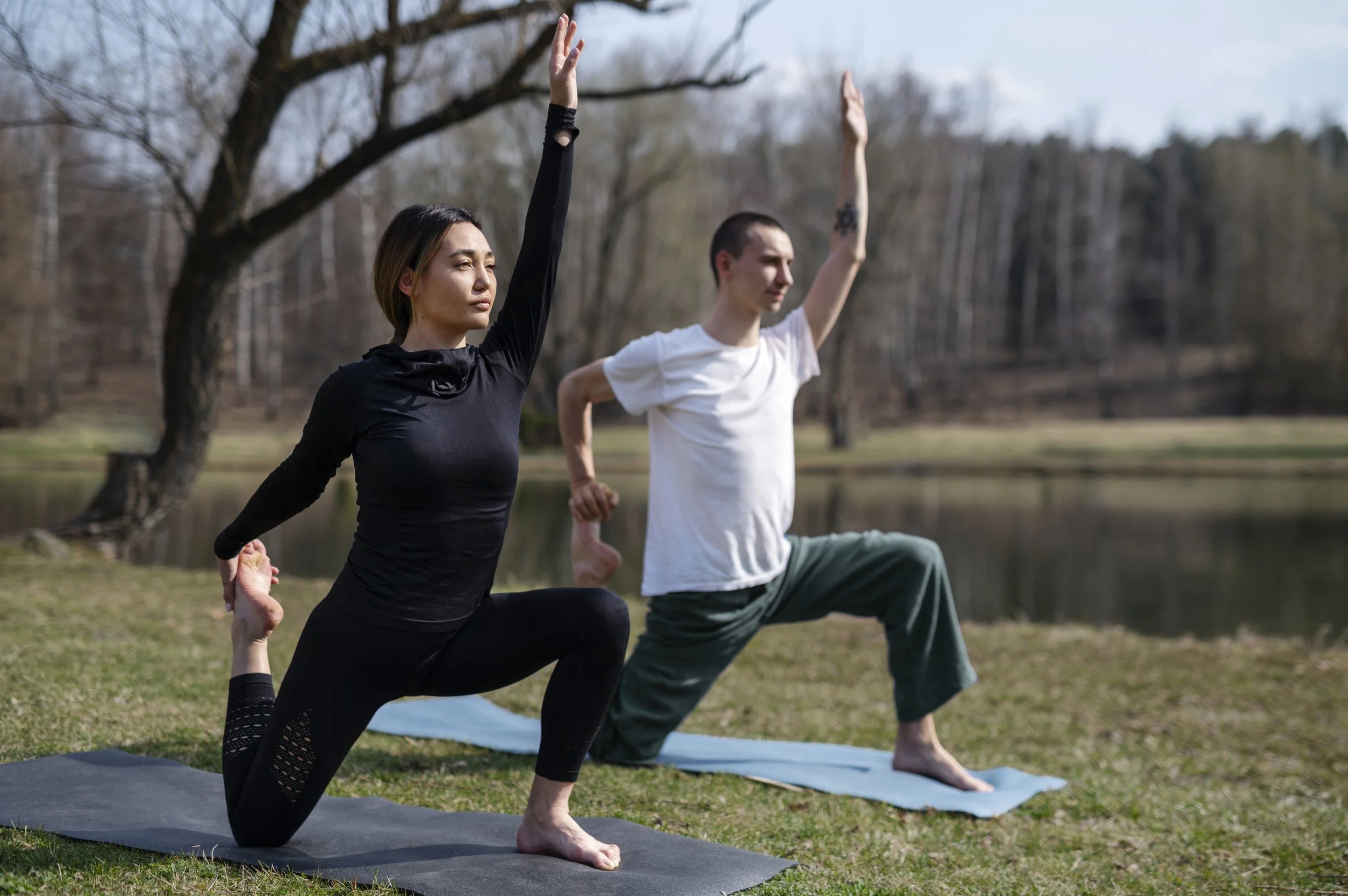 Two people practicing yoga outdoors near a lake, one woman and one man, both in a lunge pose with one arm raised and the other behind their back, on yoga mats.