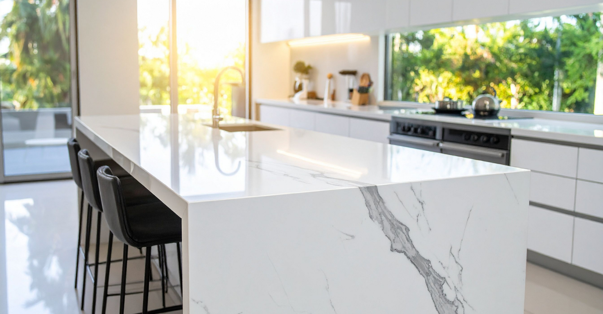 Modern kitchen with white marble island, black stools, and large windows showing green trees outside