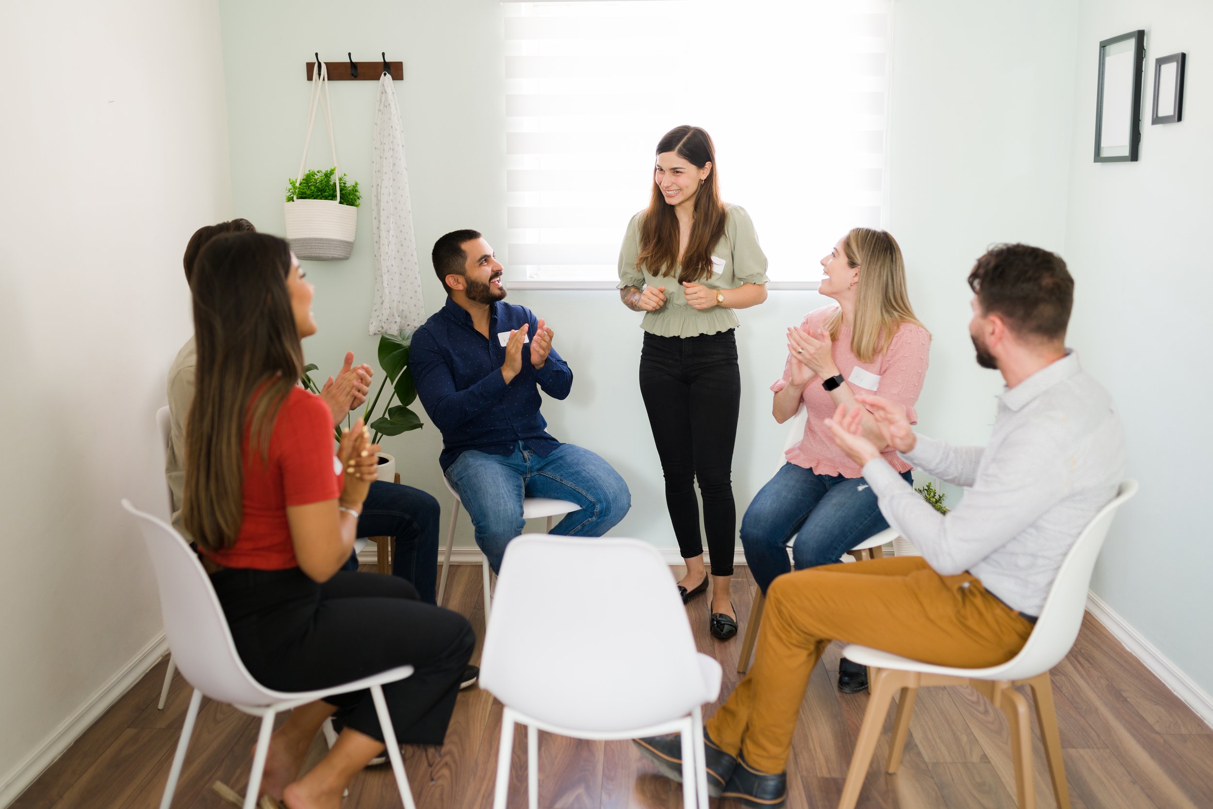 A group of six diverse people sitting in a circle in a bright room, clapping and smiling while a woman stands in the center, clapping and smiling.